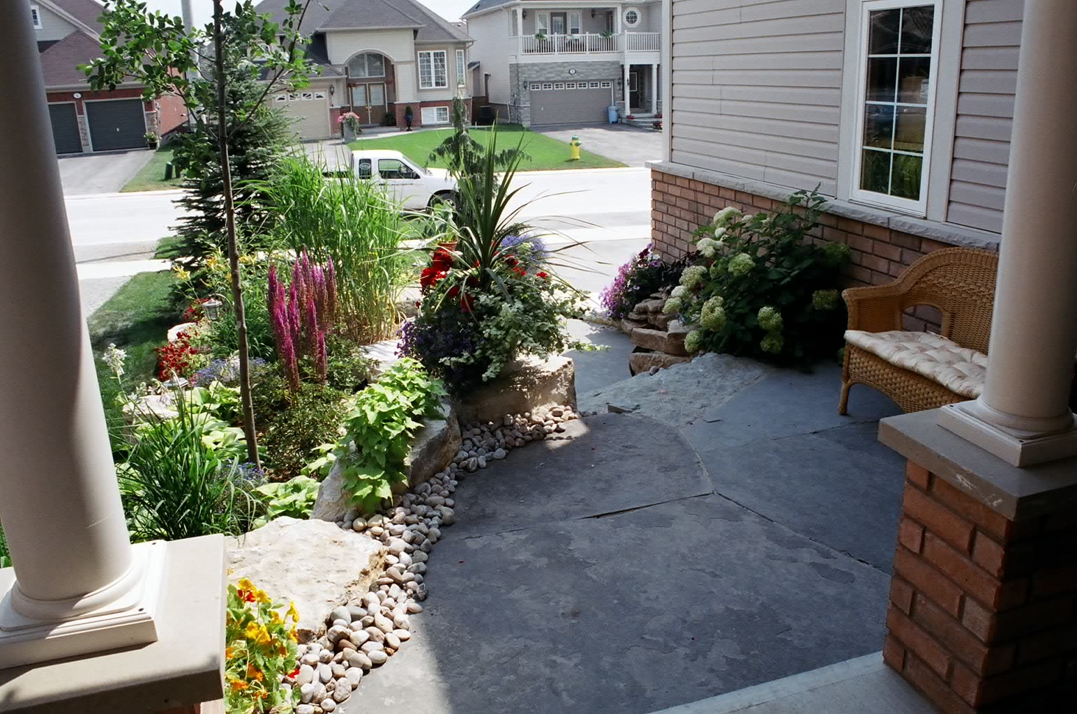 A residential front yard with a garden of diverse plants, a stone path, and wicker chair. Suburban street and houses visible.