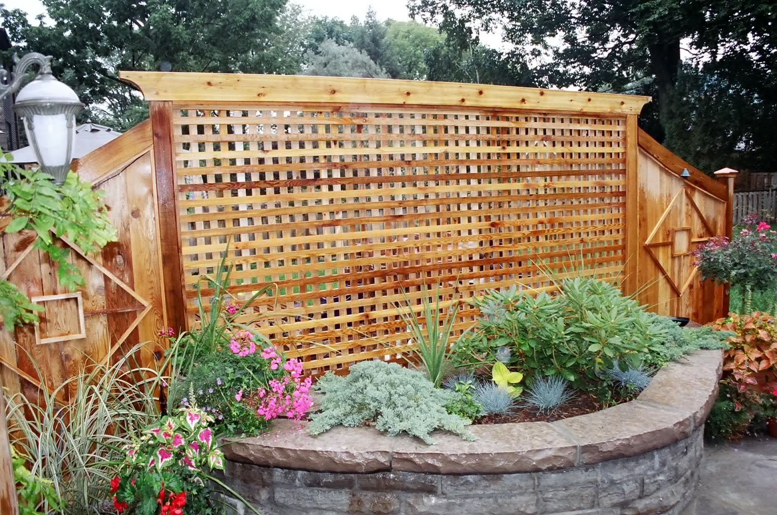 A wooden lattice fence in a garden with colorful flowers and plants, bordered by a stone wall. Lamp post visible on the side.