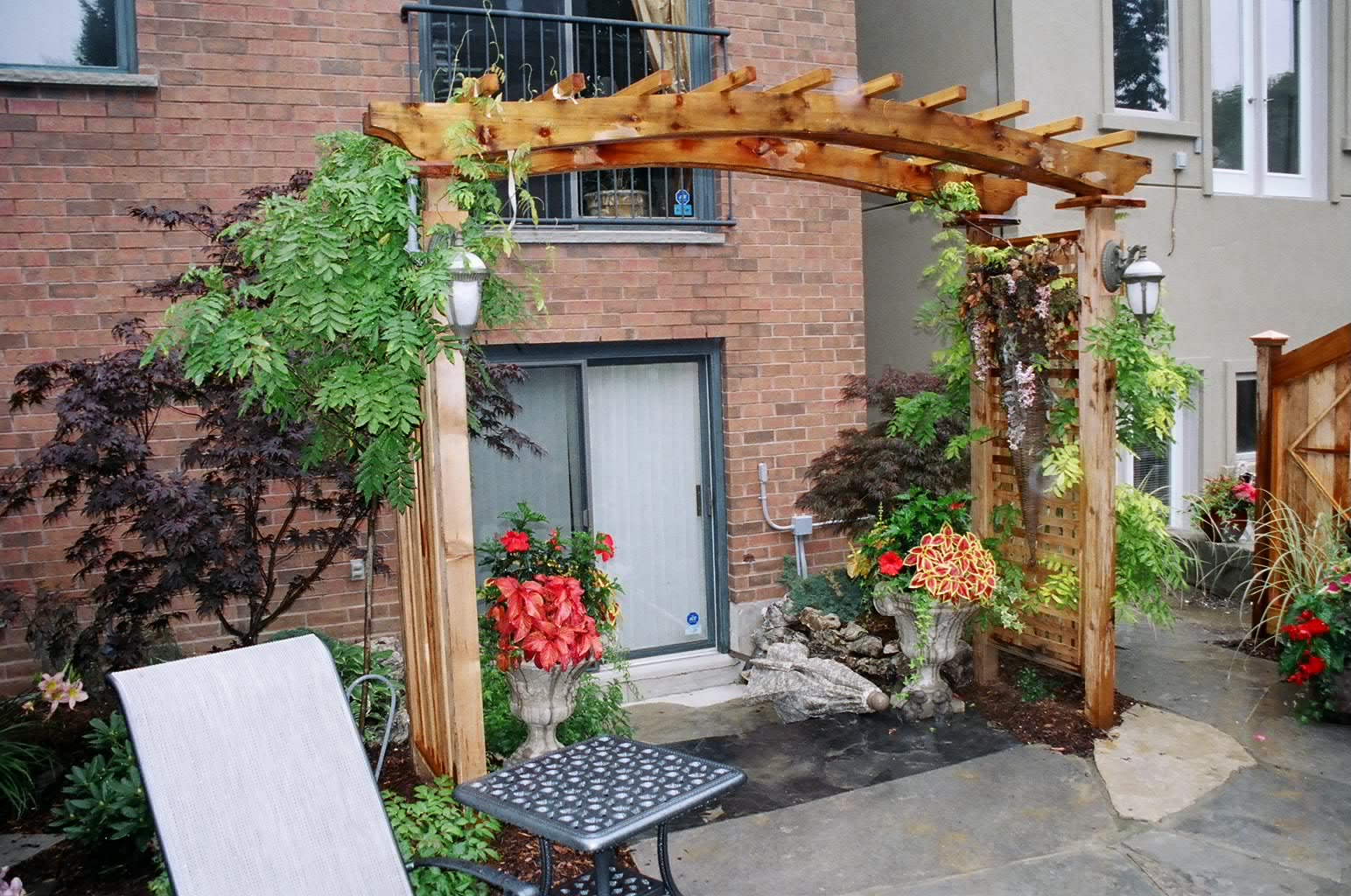 A backyard features a wooden pergola, lush plants, and a small patio set against a brick house with a glass door.