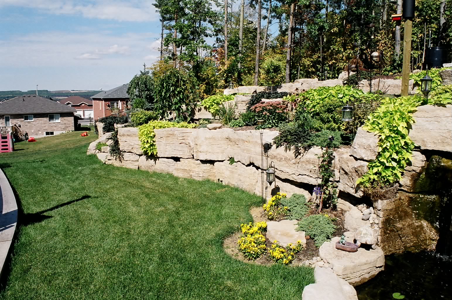 The image shows a landscaped backyard garden with stone walls, plants, and flowers. Residential houses and trees are visible in the background.