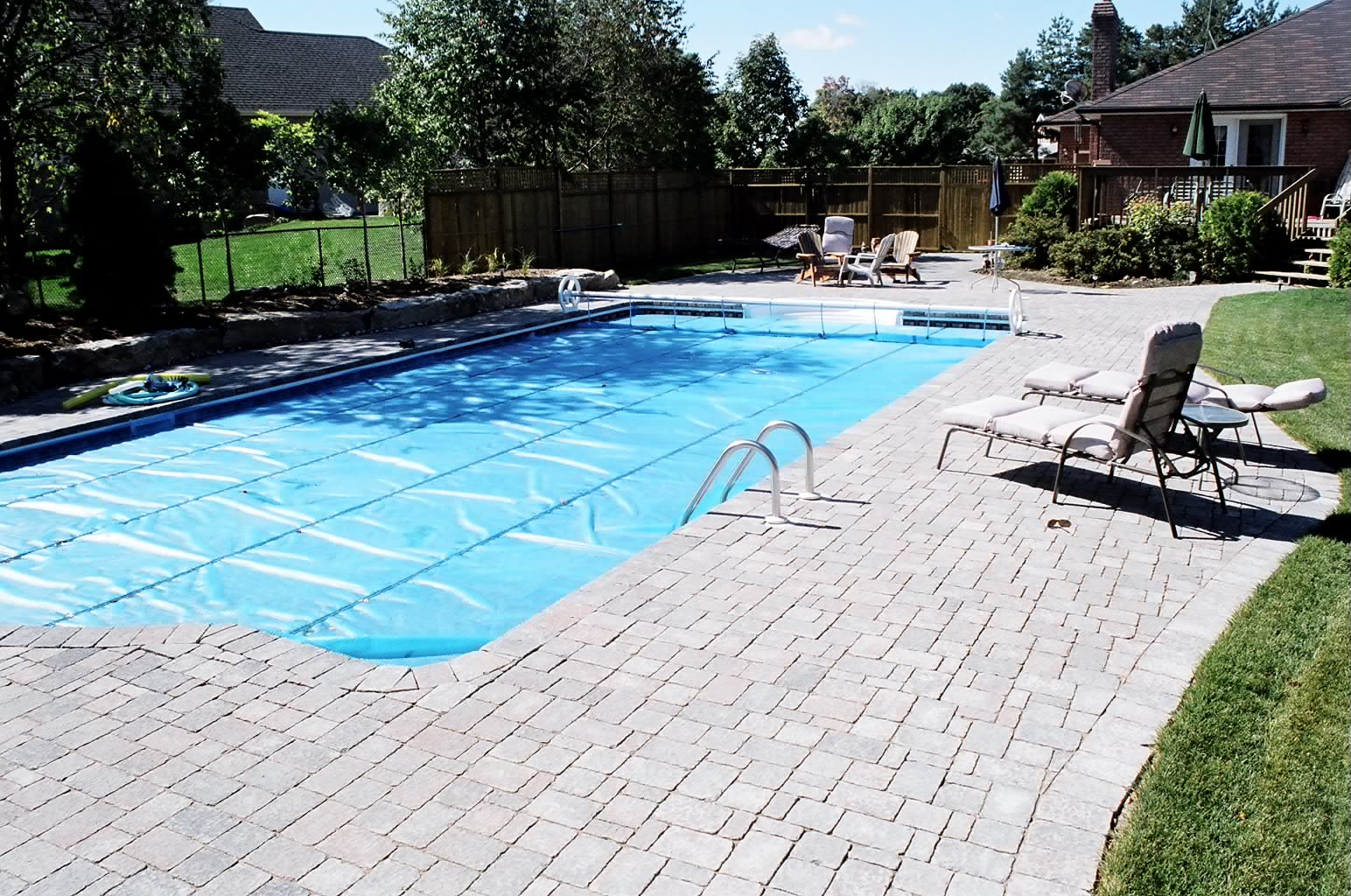 A backyard features a rectangular swimming pool, chairs, and green lawn, enclosed by a wooden fence and surrounded by trees and a house.