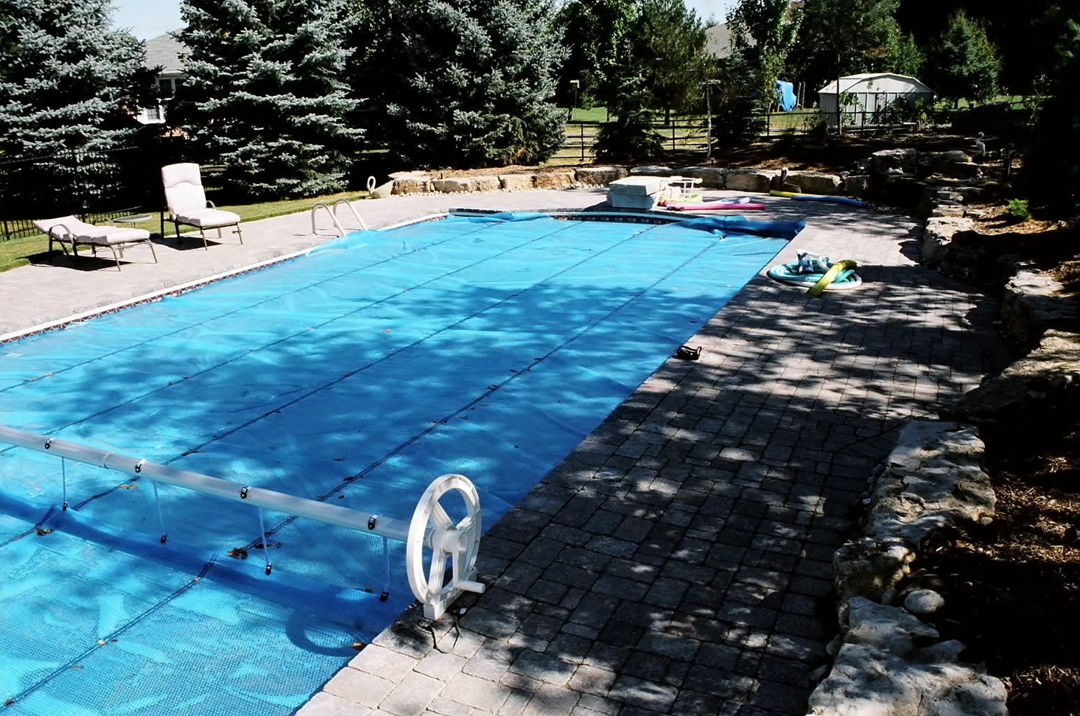 A covered rectangular swimming pool surrounded by trees and stone, with lounge chairs and pool toys, on a sunny day.