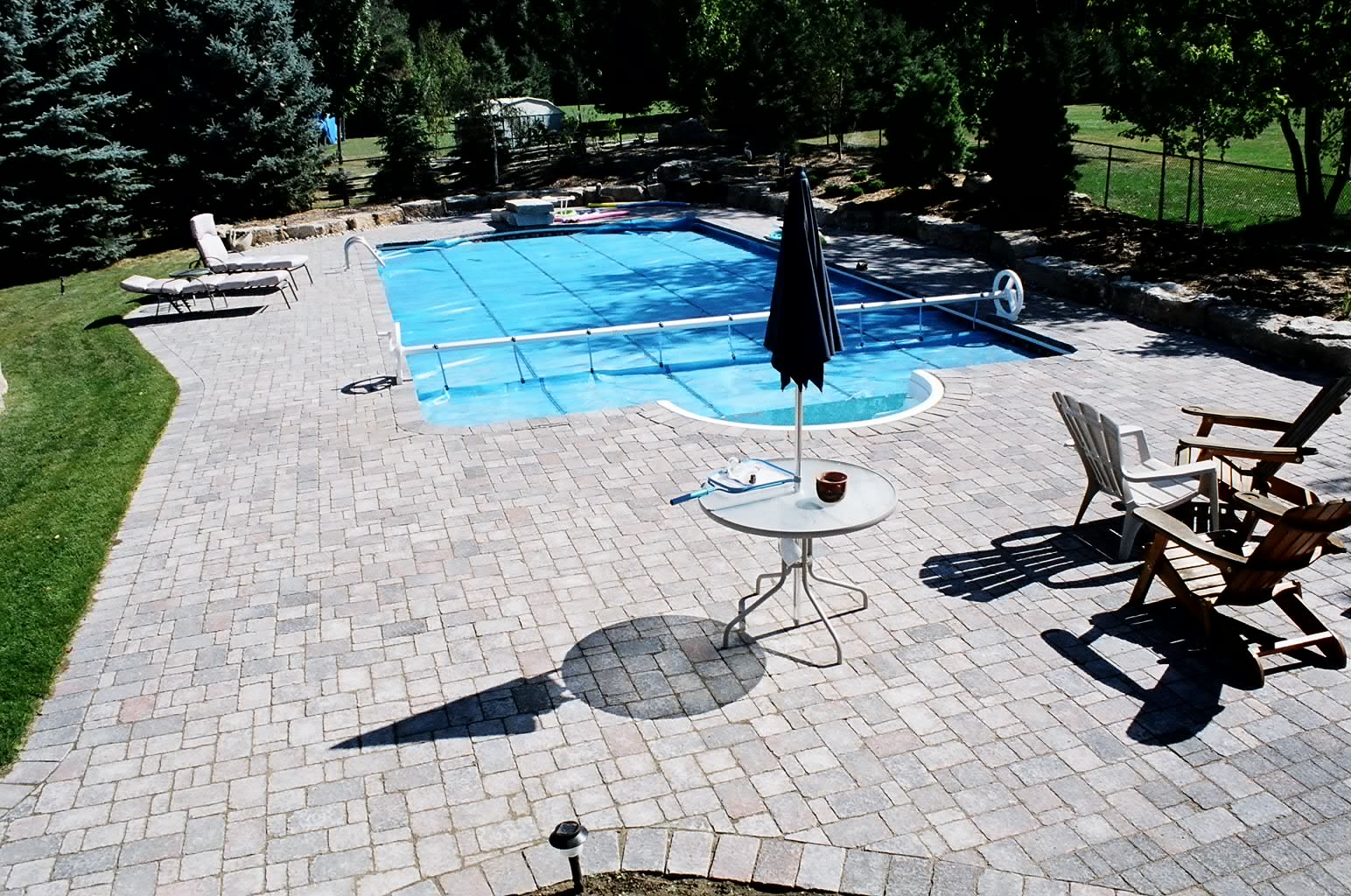 Backyard with a rectangular pool covered, surrounded by pavers. Trees border the area, and patio furniture includes a table and chairs.