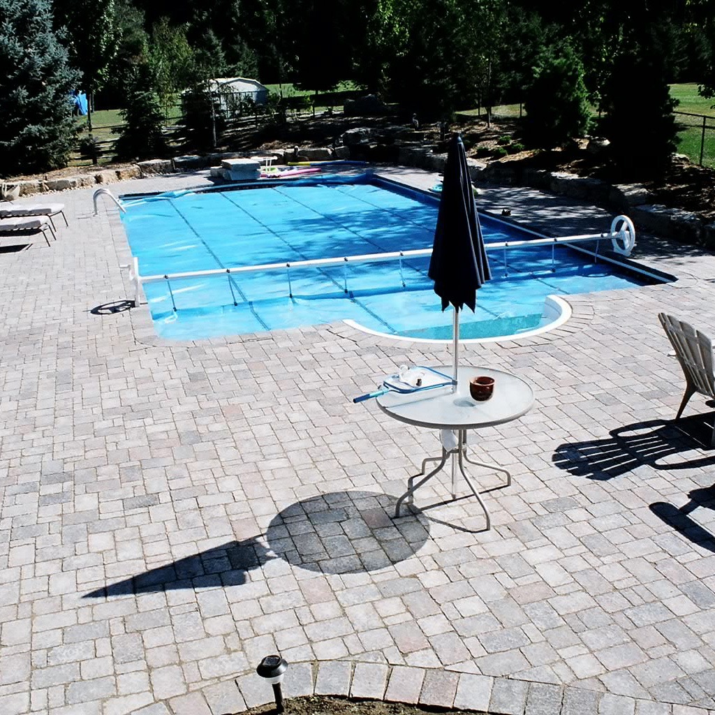 A spacious outdoor area with a swimming pool, surrounded by trees. Patio furniture and a table with an umbrella are nearby.