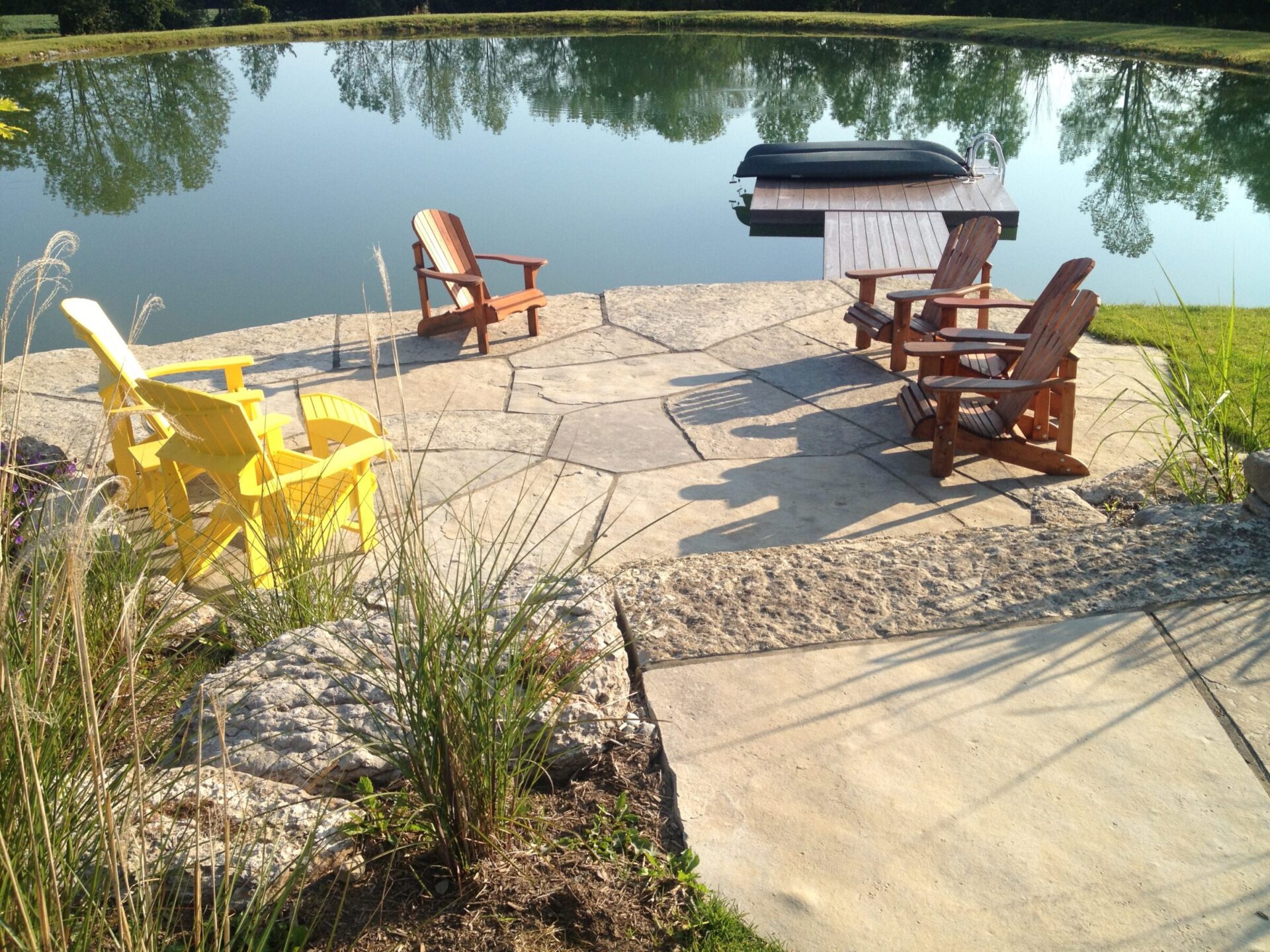 A stone patio with Adirondack chairs overlooks a serene pond, surrounded by greenery and featuring a small dock with a boat.