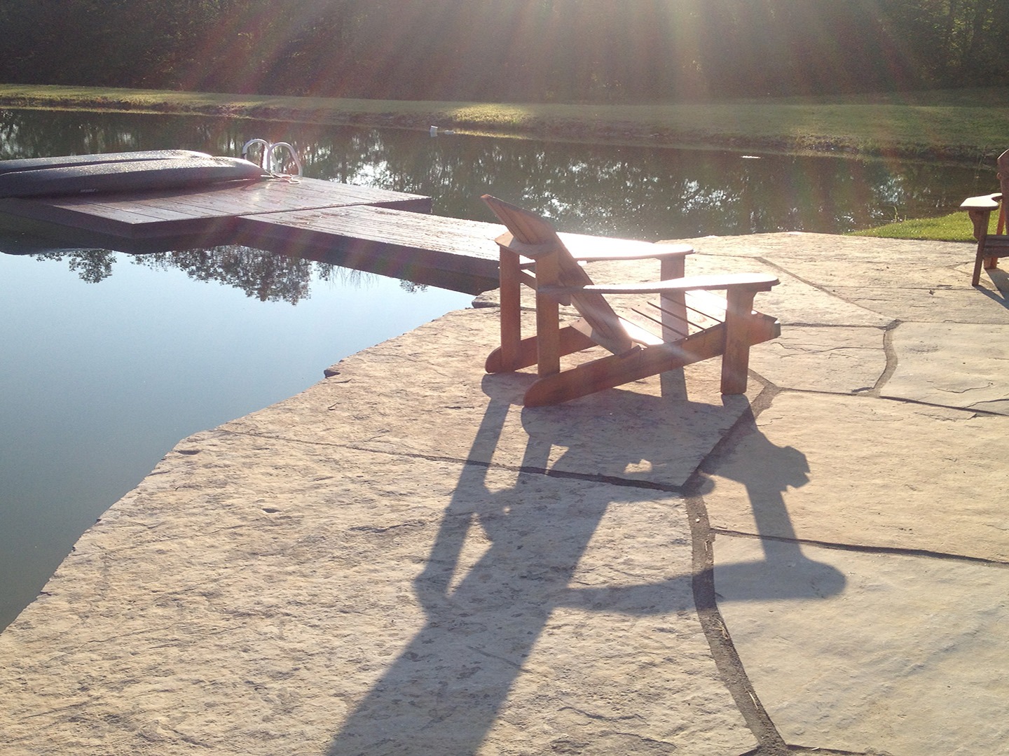 Stone patio with wooden chairs overlooking a calm pond. Sunlight creates long shadows across the ground, adding a serene, tranquil atmosphere.