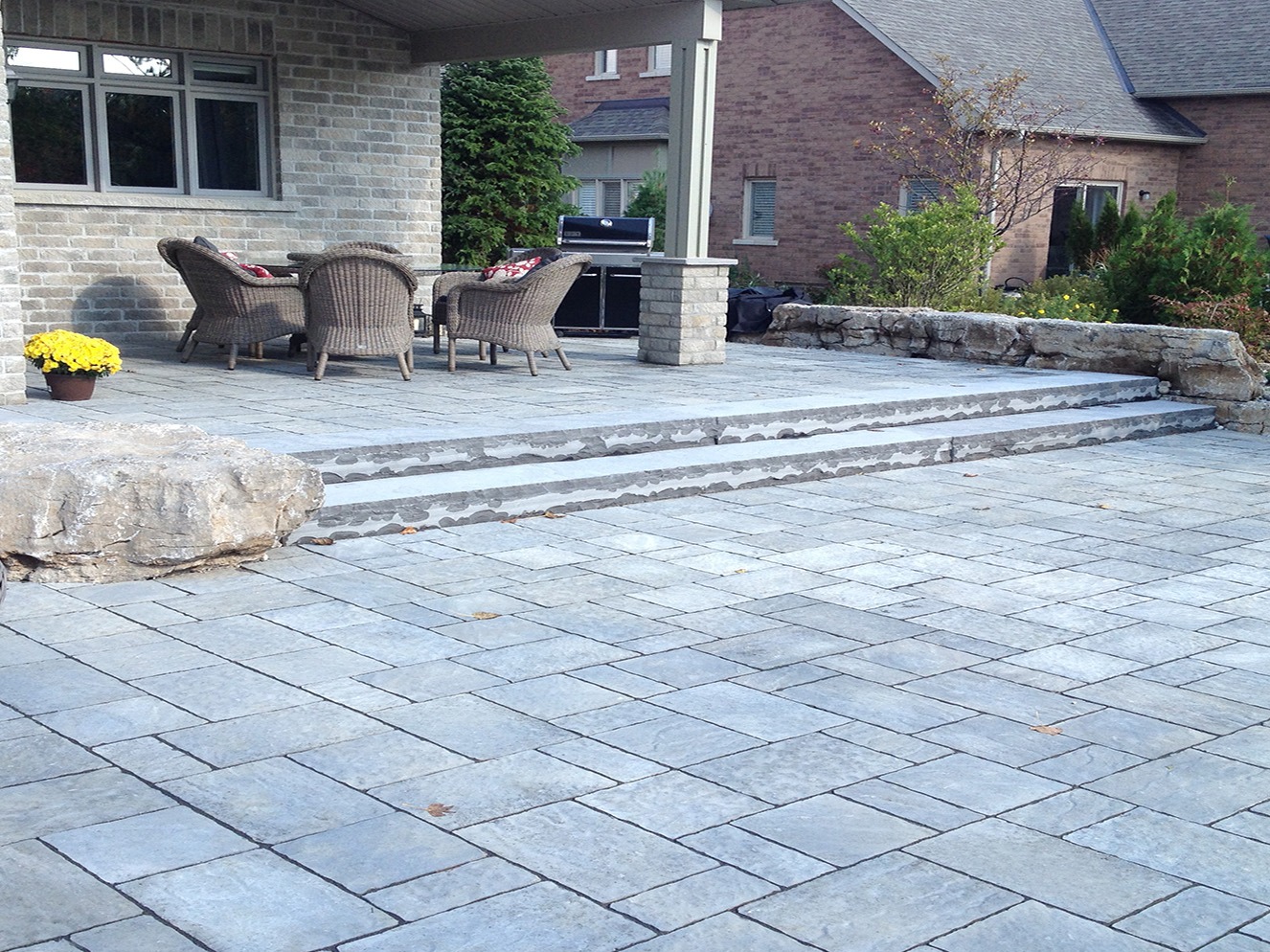 A spacious stone patio with wicker furniture and potted flowers, adjacent to a brick house and surrounded by a natural stone retaining wall.