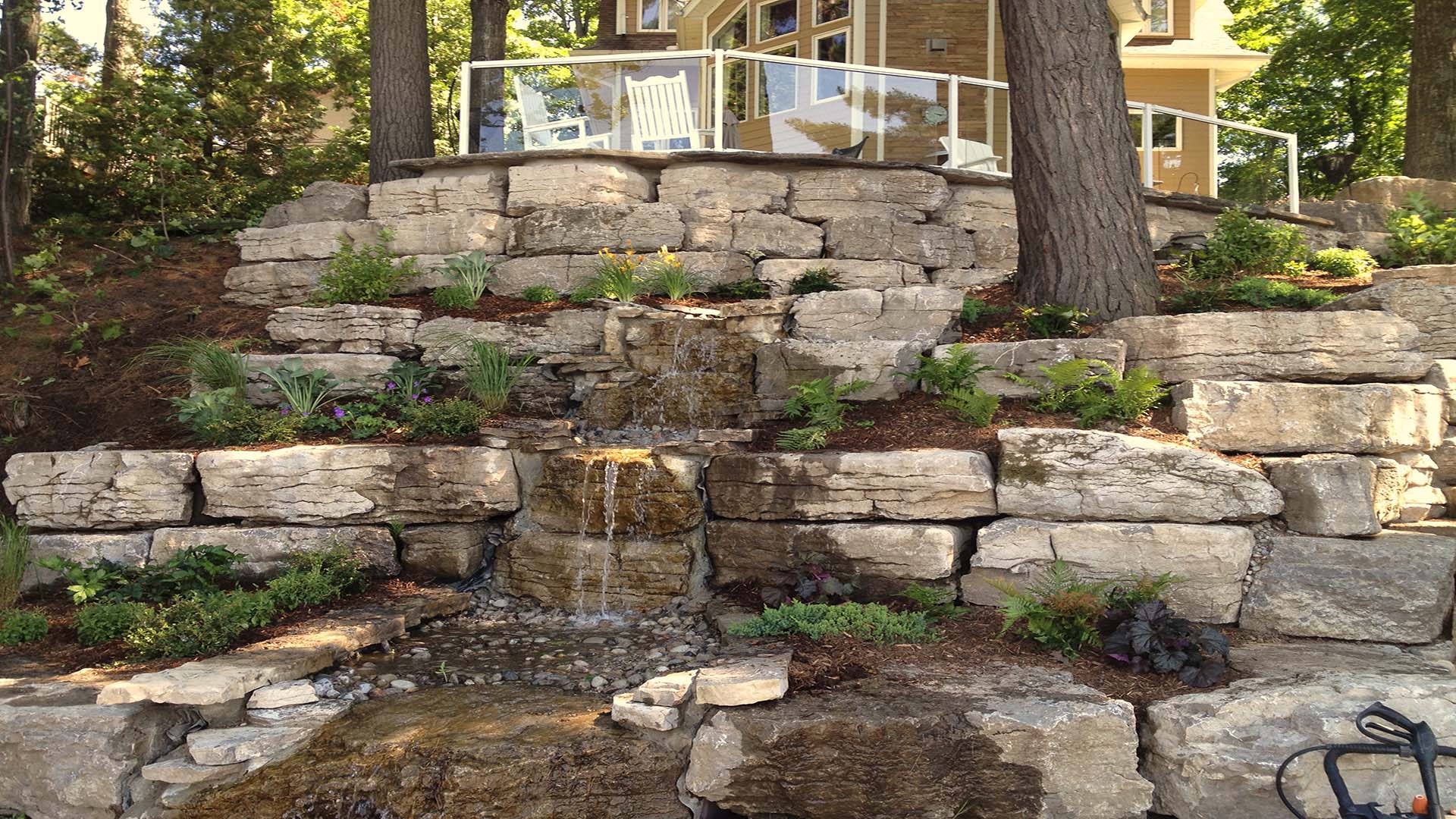 A beautifully landscaped stone terrace features a small cascading waterfall, surrounded by lush greenery. A modern house is visible above, behind glass railings.