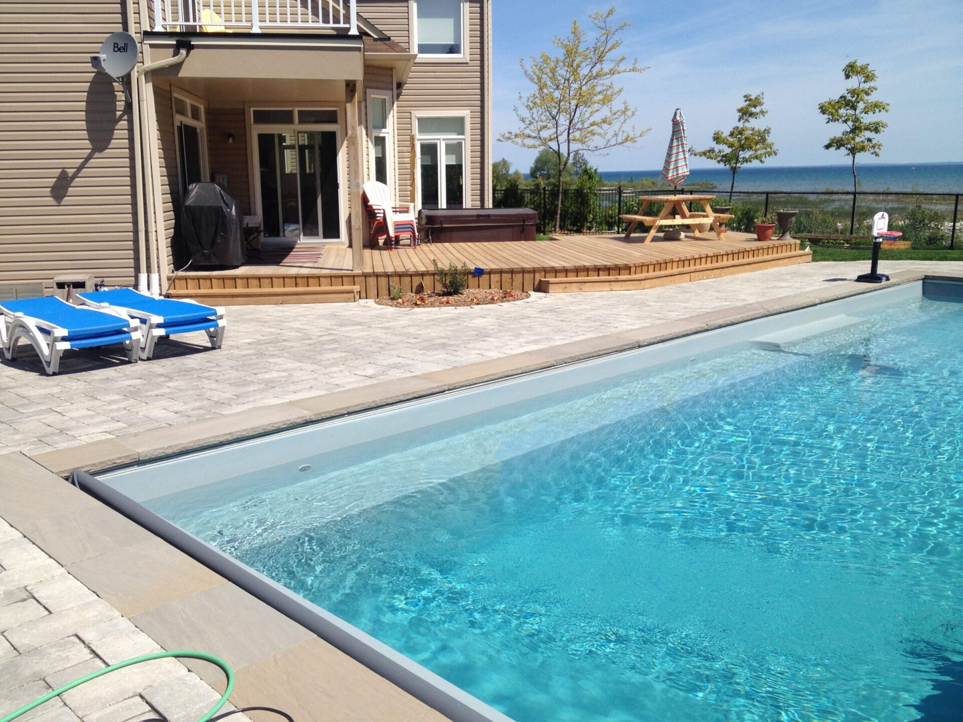 A backyard pool with loungers, a deck, and a clear sky. The view overlooks a grassy area and a body of water beyond.