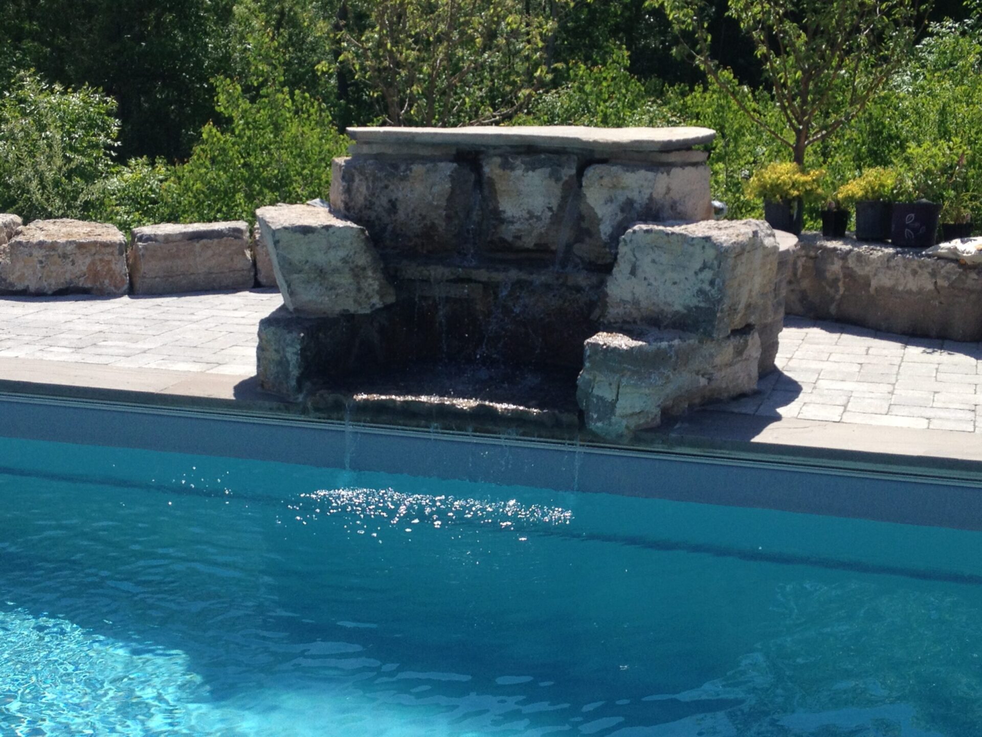 Stone waterfall feature flows into a clear blue swimming pool. Surrounded by greenery and stone paving, creating a tranquil backyard setting.