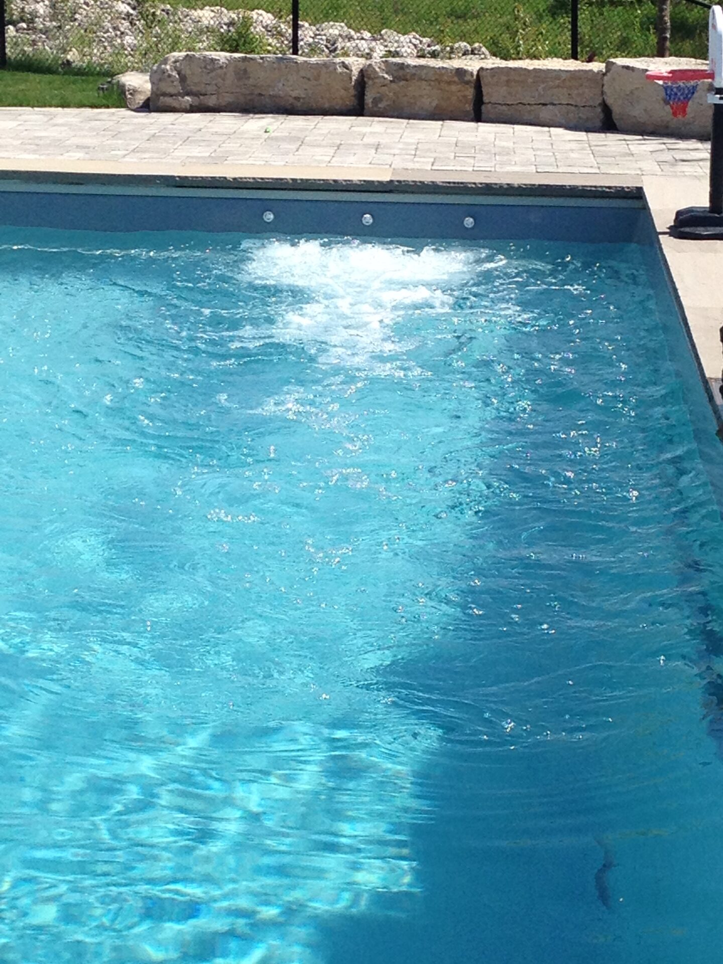 A clear pool with bubbling water, surrounded by stone patio and rocks. A basketball hoop is visible near the water's edge.