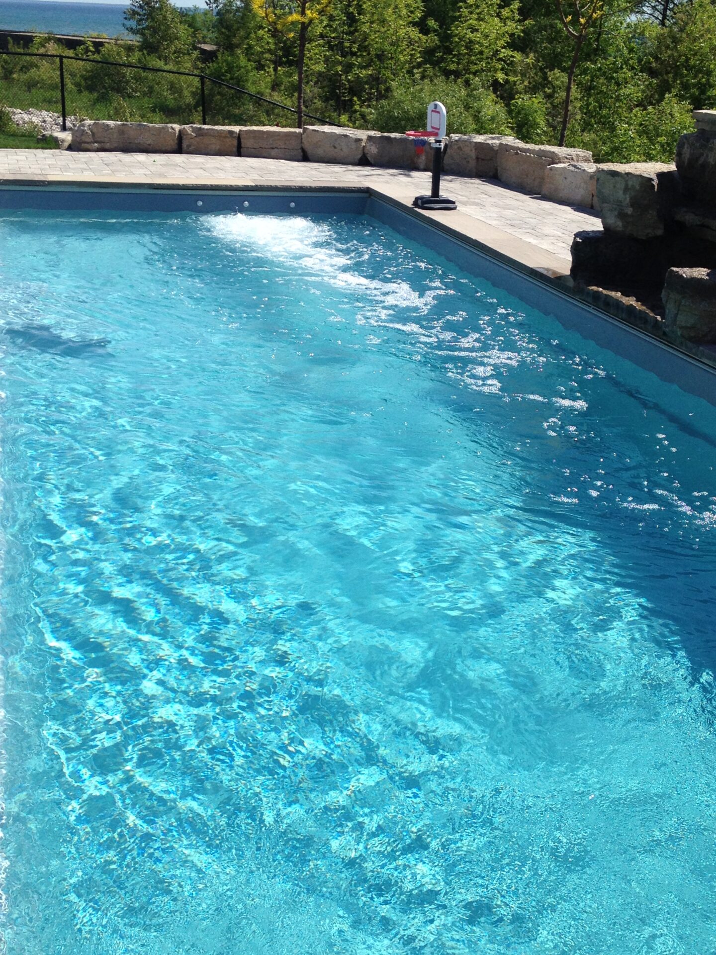 A swimming pool with clear blue water, surrounded by stone tiles and rock edges, overlooks a lush wooded area under a sunny sky.