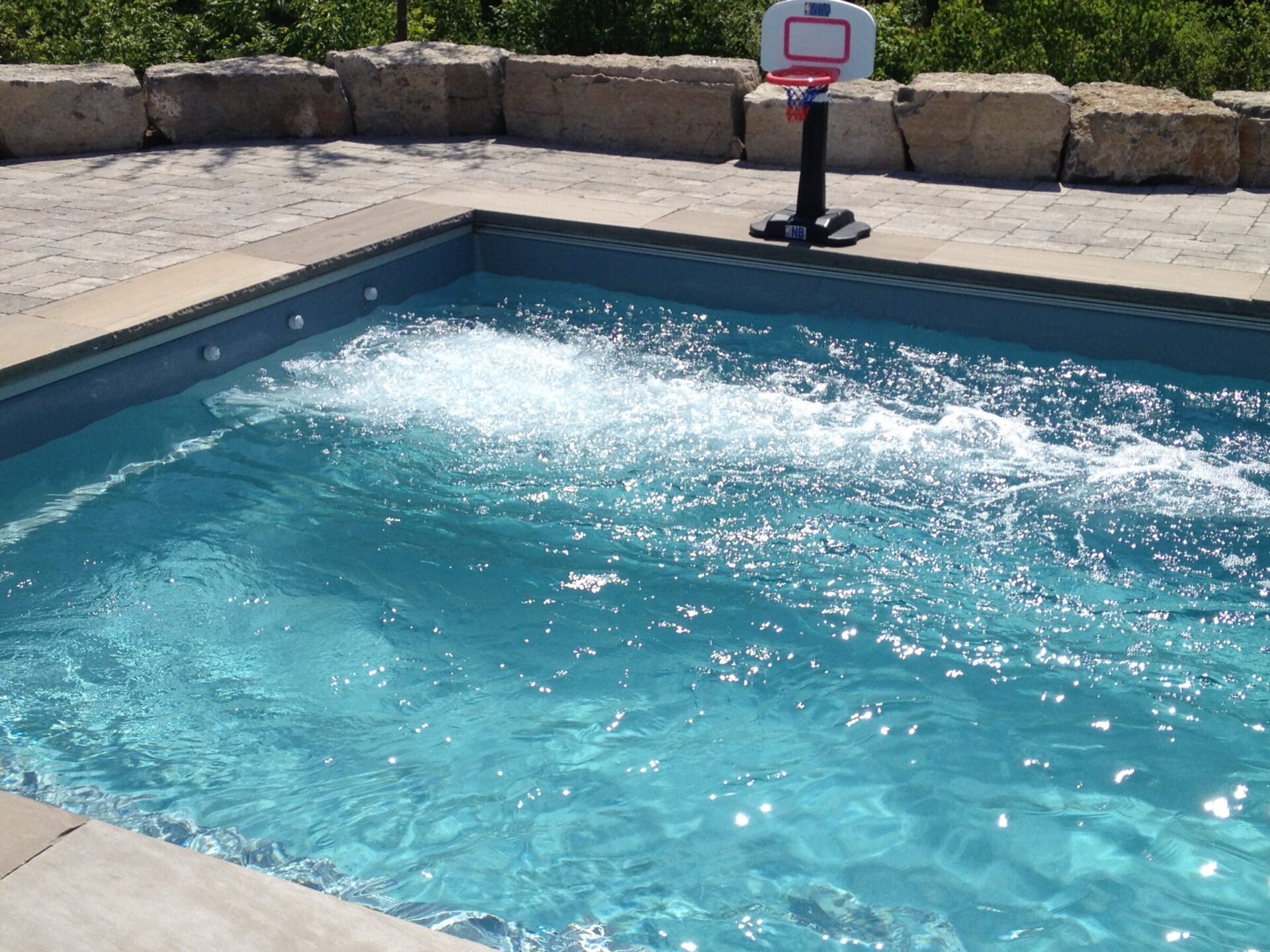 A clear swimming pool with bubbling water features a basketball hoop nearby, surrounded by stone landscaping and sunlight.