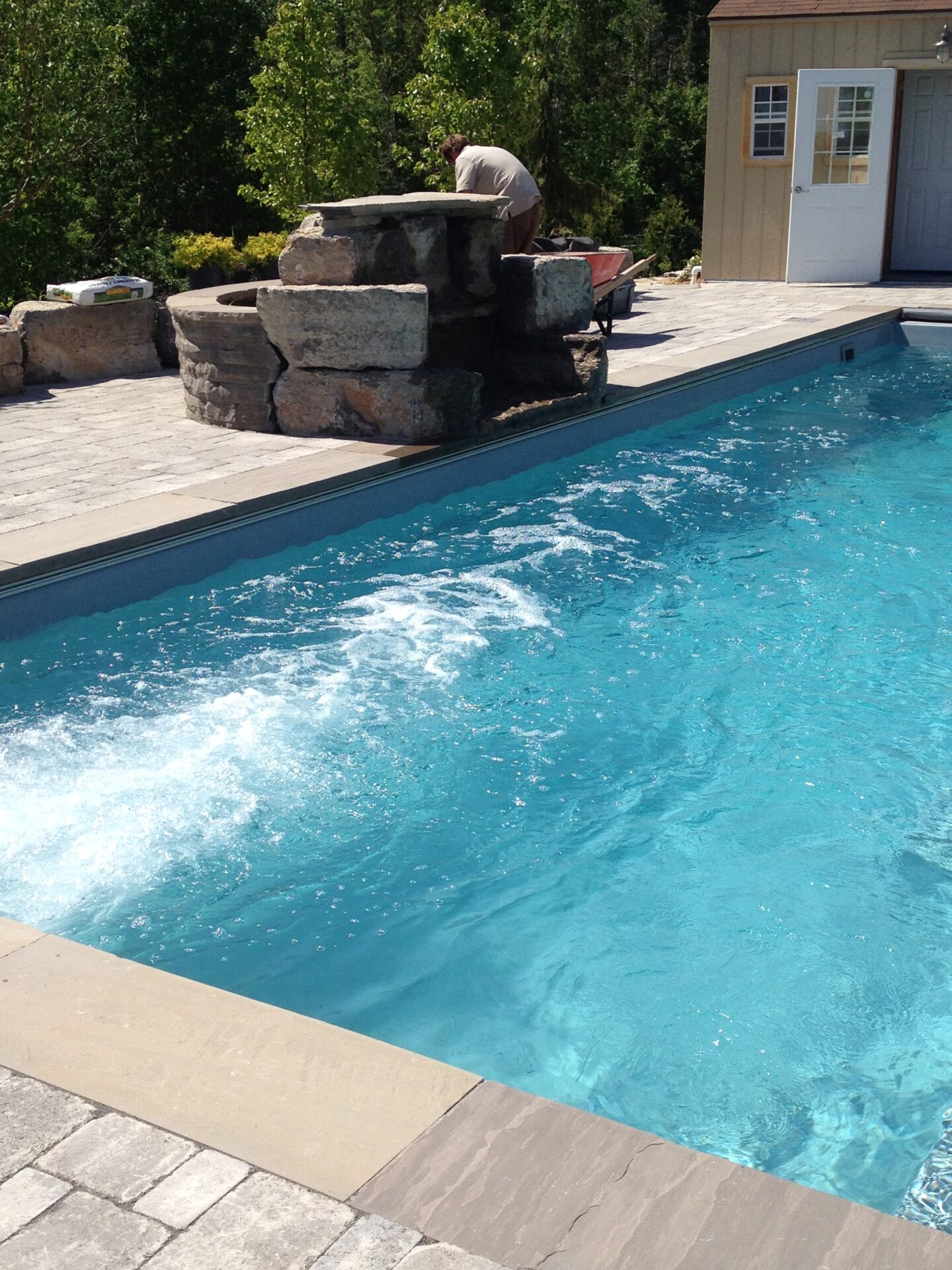 A person works near a blue swimming pool with a rock waterfall feature and a small shed, surrounded by trees.