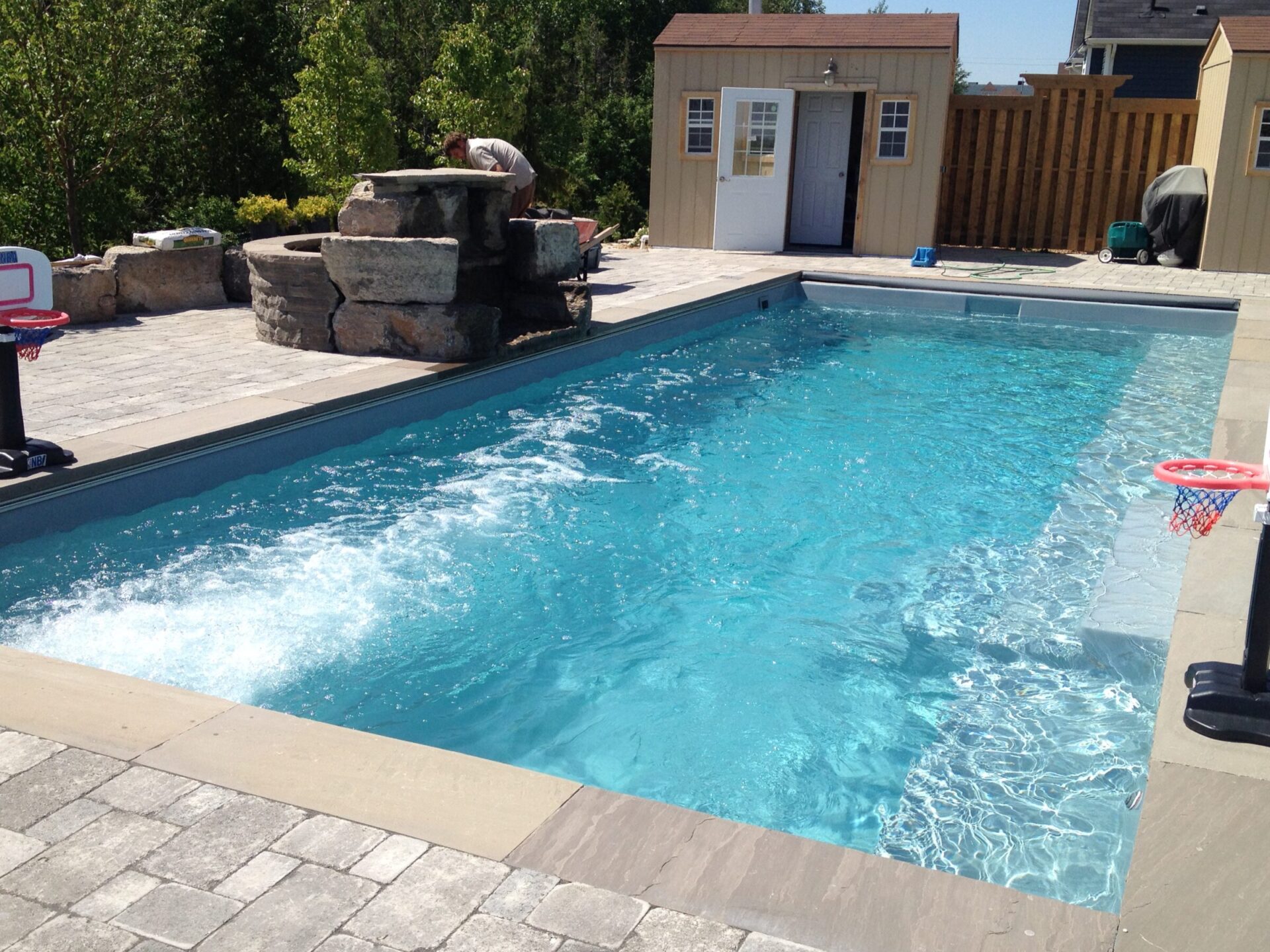 A backyard features a rectangular pool, stone waterfall, basketball hoops, and two sheds. A person tends to the waterfall.