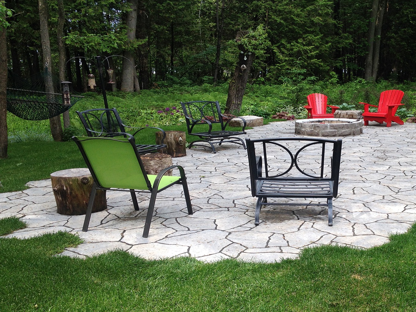 A peaceful outdoor patio with green and red chairs, a stone fire pit, surrounded by lush greenery and trees, creates a relaxing atmosphere.