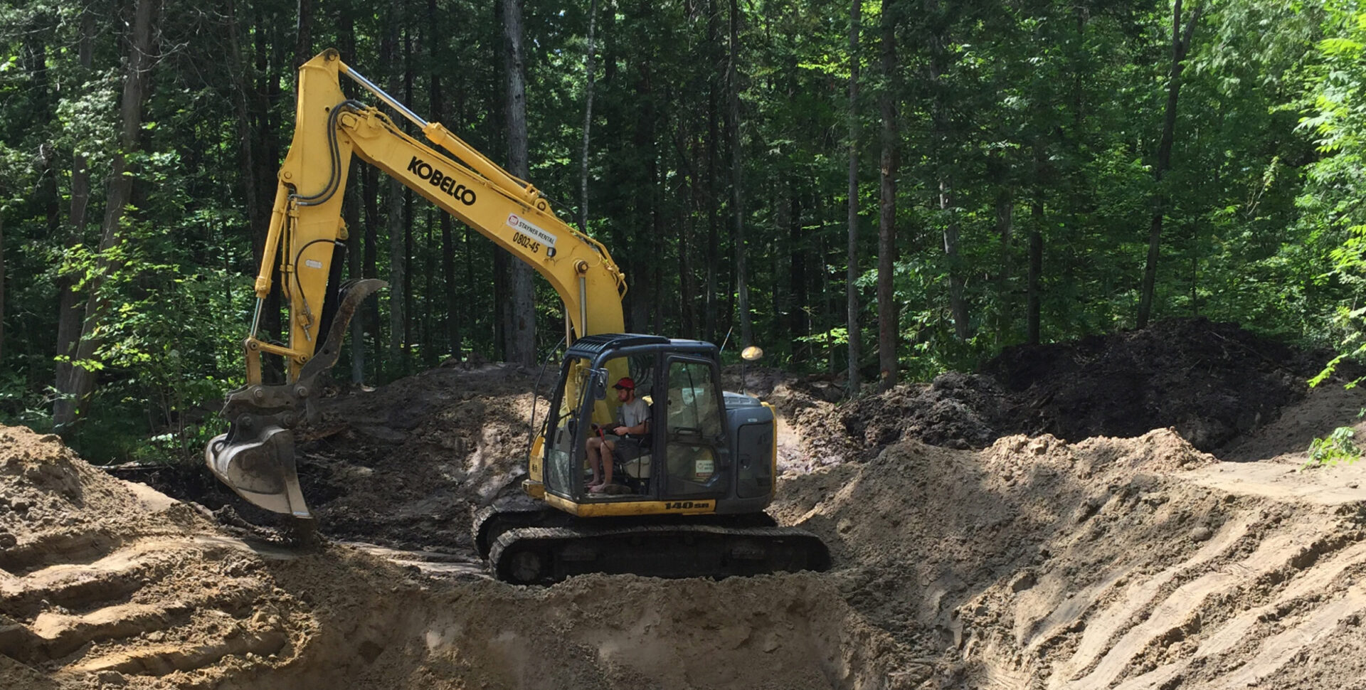 An excavator operates in a forested area, digging around a waterlogged pit, surrounded by piles of earth and dense green foliage.