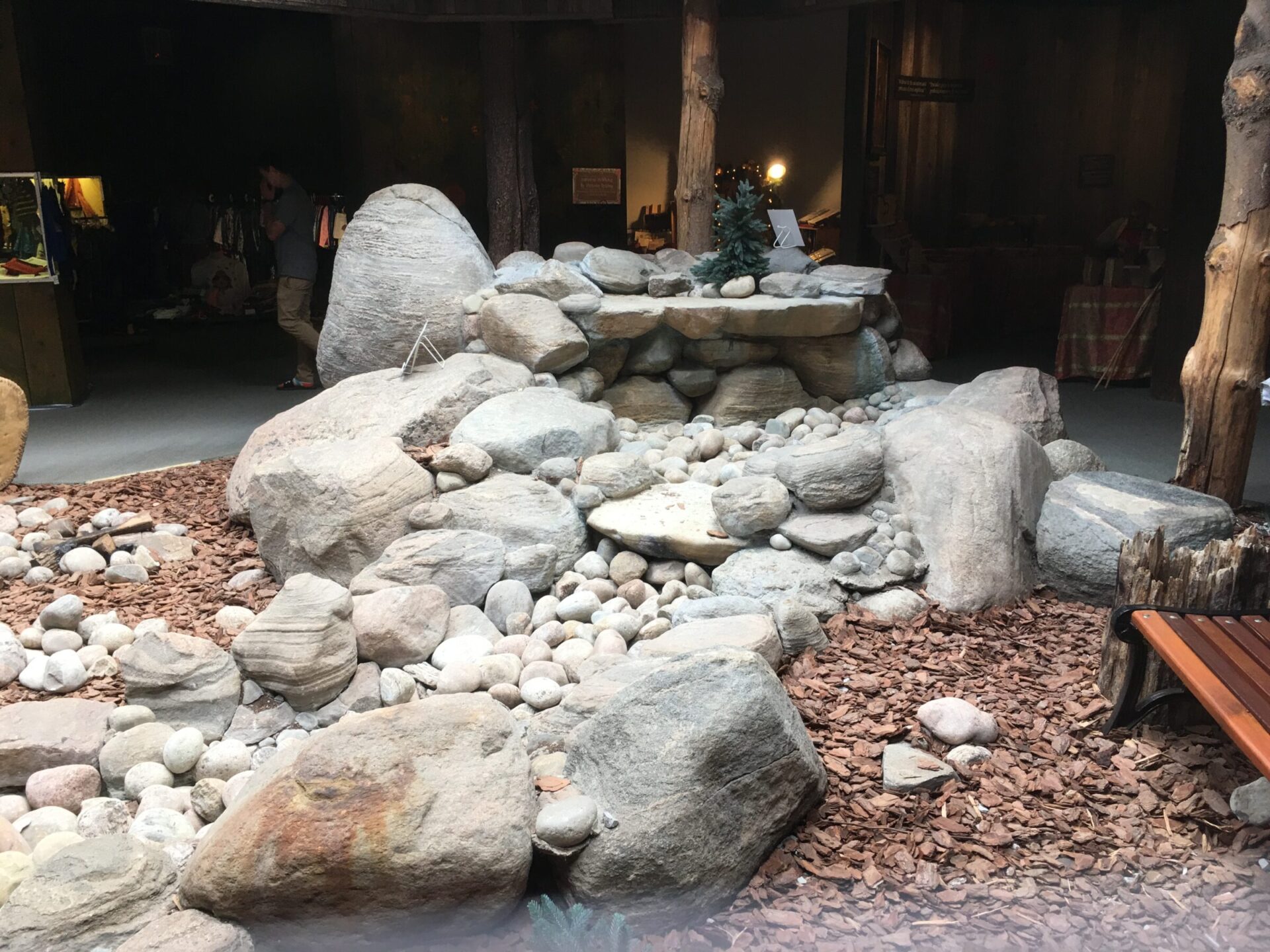 Rocky indoor garden display with assorted stones, small tree, and surrounding wooden elements. Dim lighting and a person standing in the background.