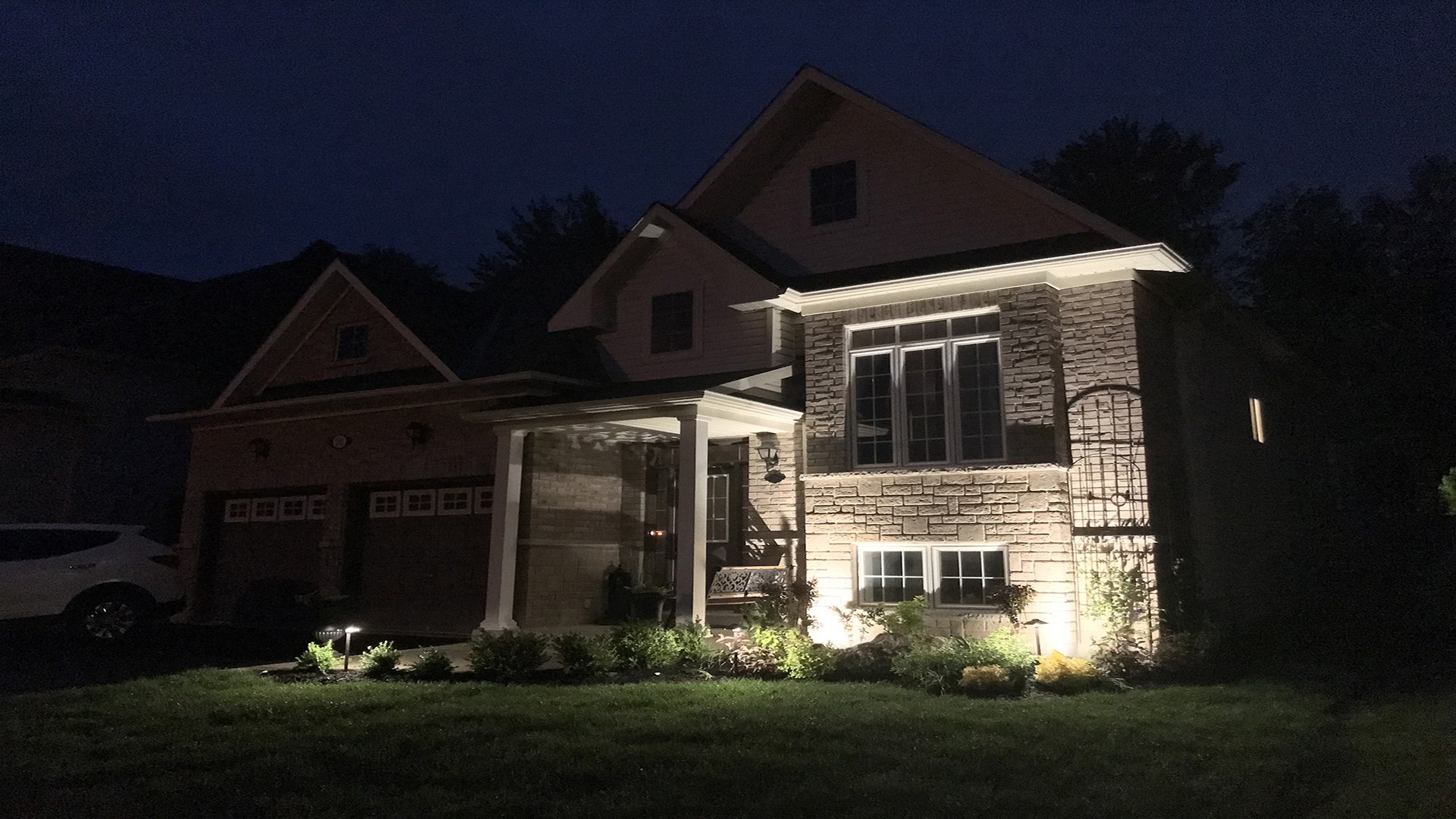 A suburban house at night, front porch illuminated. Windows and garden are visible, with a car parked in the driveway.