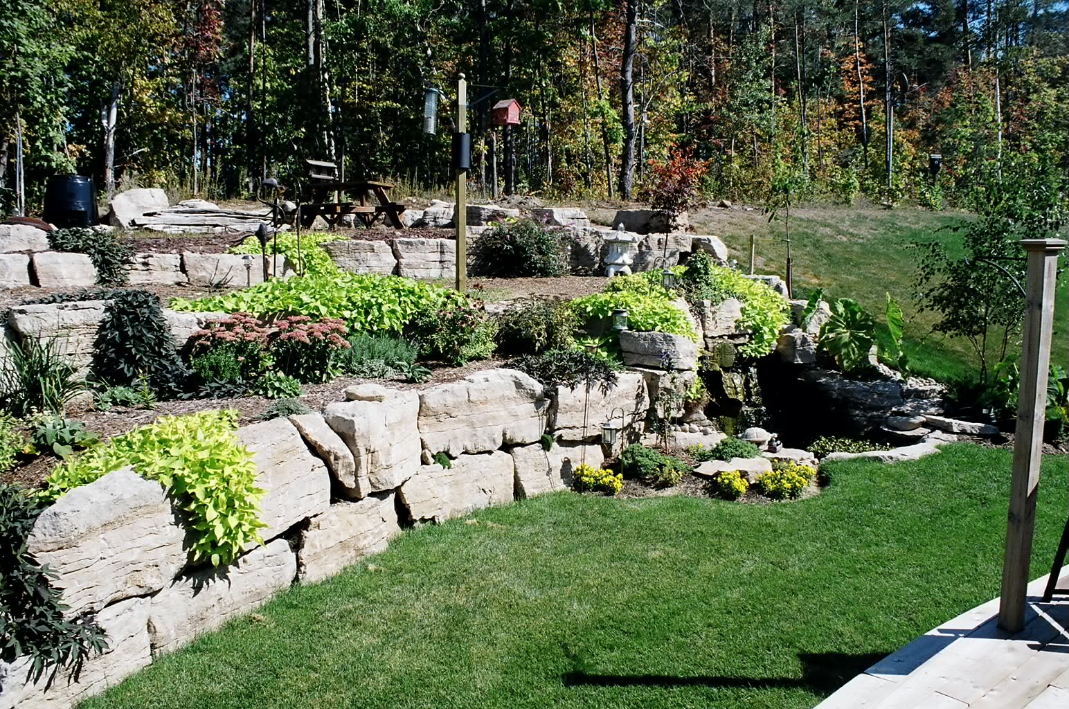 Terraced garden with stone walls, lush greenery, small waterfall, and pathway. Surrounded by dense forest, creating a serene, natural environment.