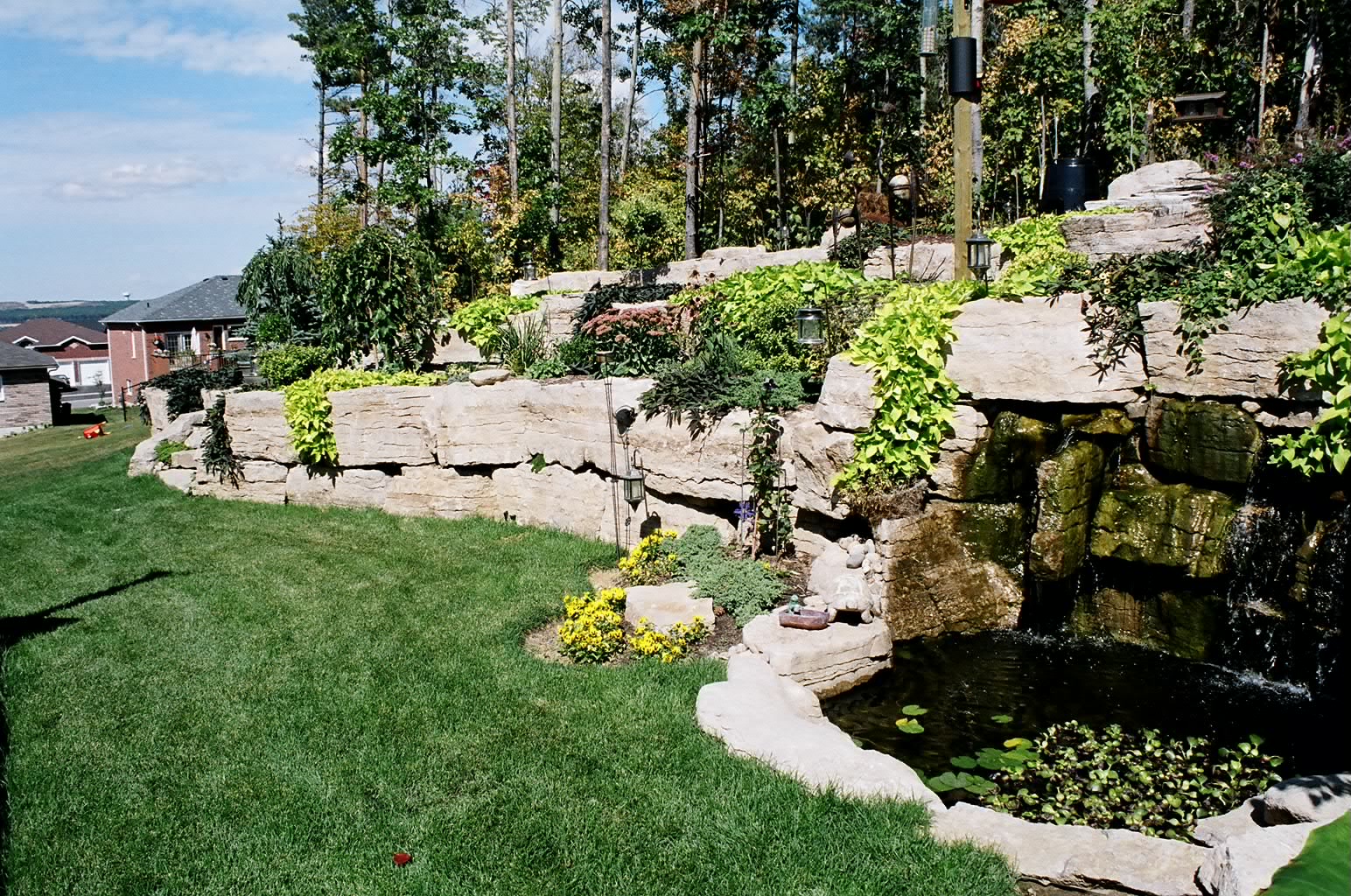 Rock garden with lush greenery, cascading waterfall, and a small pond. Surrounded by grass, trees, and distant residential homes under a blue sky.