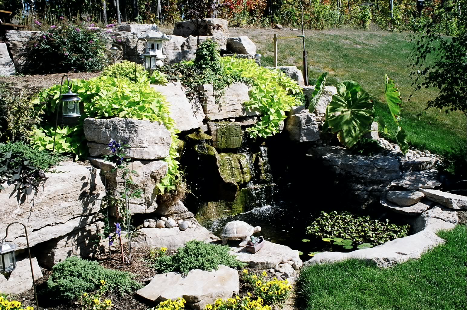 A serene garden scene features natural rock formations, cascading water, lush greenery, and a small pond, creating a peaceful outdoor sanctuary.