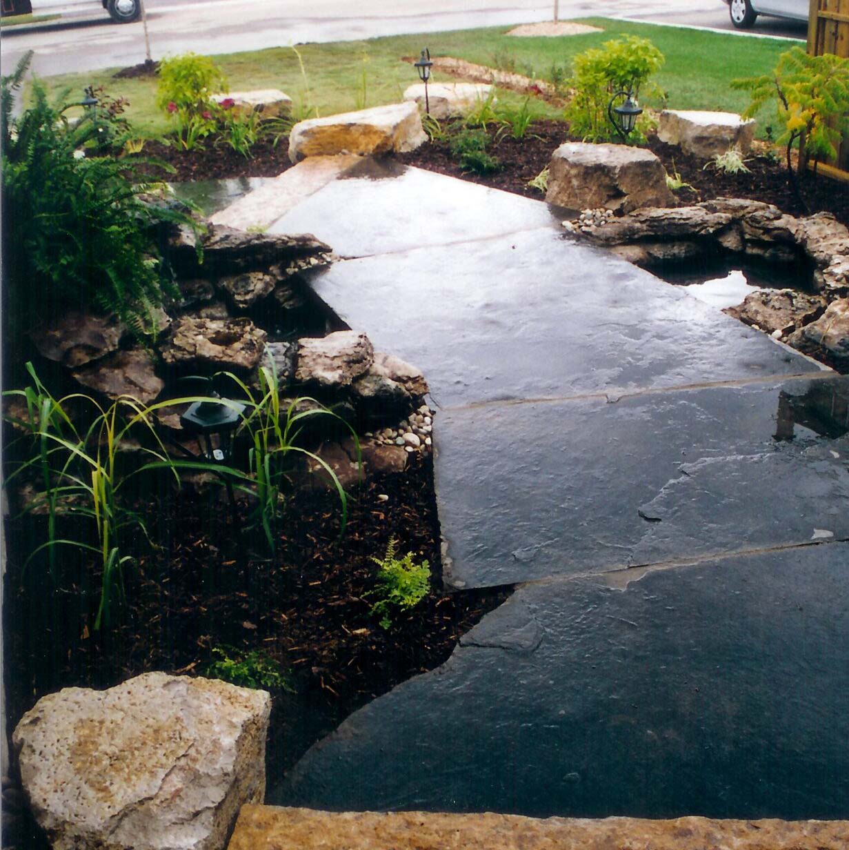 A serene garden path with large stepping stones, bordered by rocks and greenery, leads through a neatly landscaped area under overcast skies.