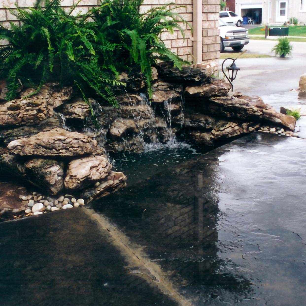 A small, rocky waterfall with surrounding ferns flows into a shallow pool on a paved area in a suburban neighborhood.