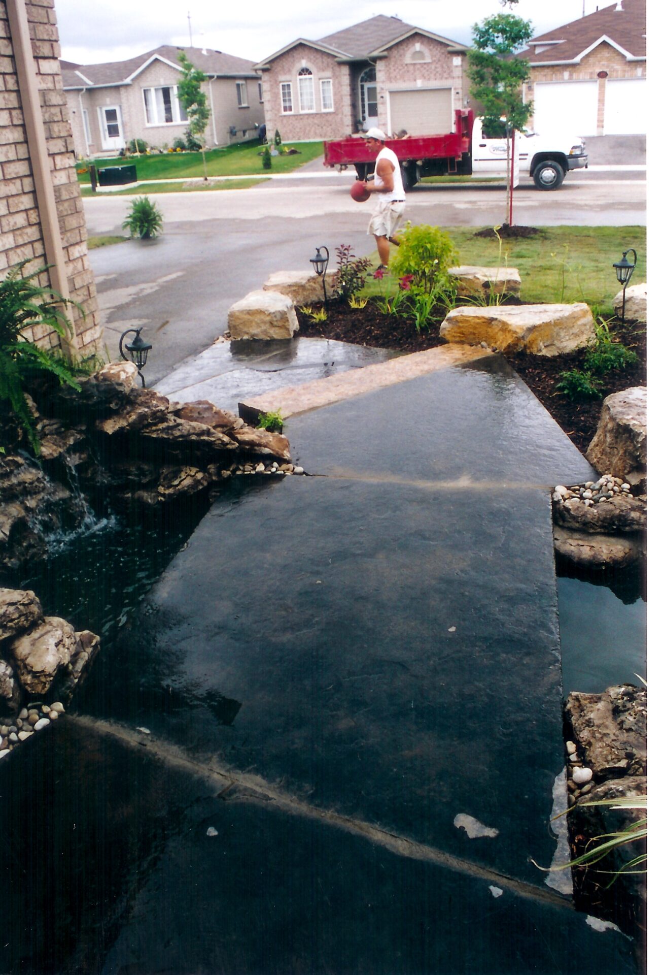 A person plays basketball in a suburban neighborhood with neatly maintained lawns, rock landscaping, and a small water feature in the foreground.