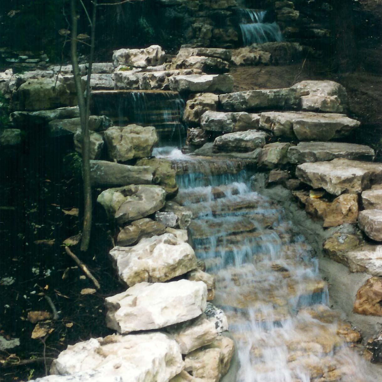 A small waterfall flows over layered rocks in a wooded area, surrounded by trees and foliage, creating a serene natural scene.