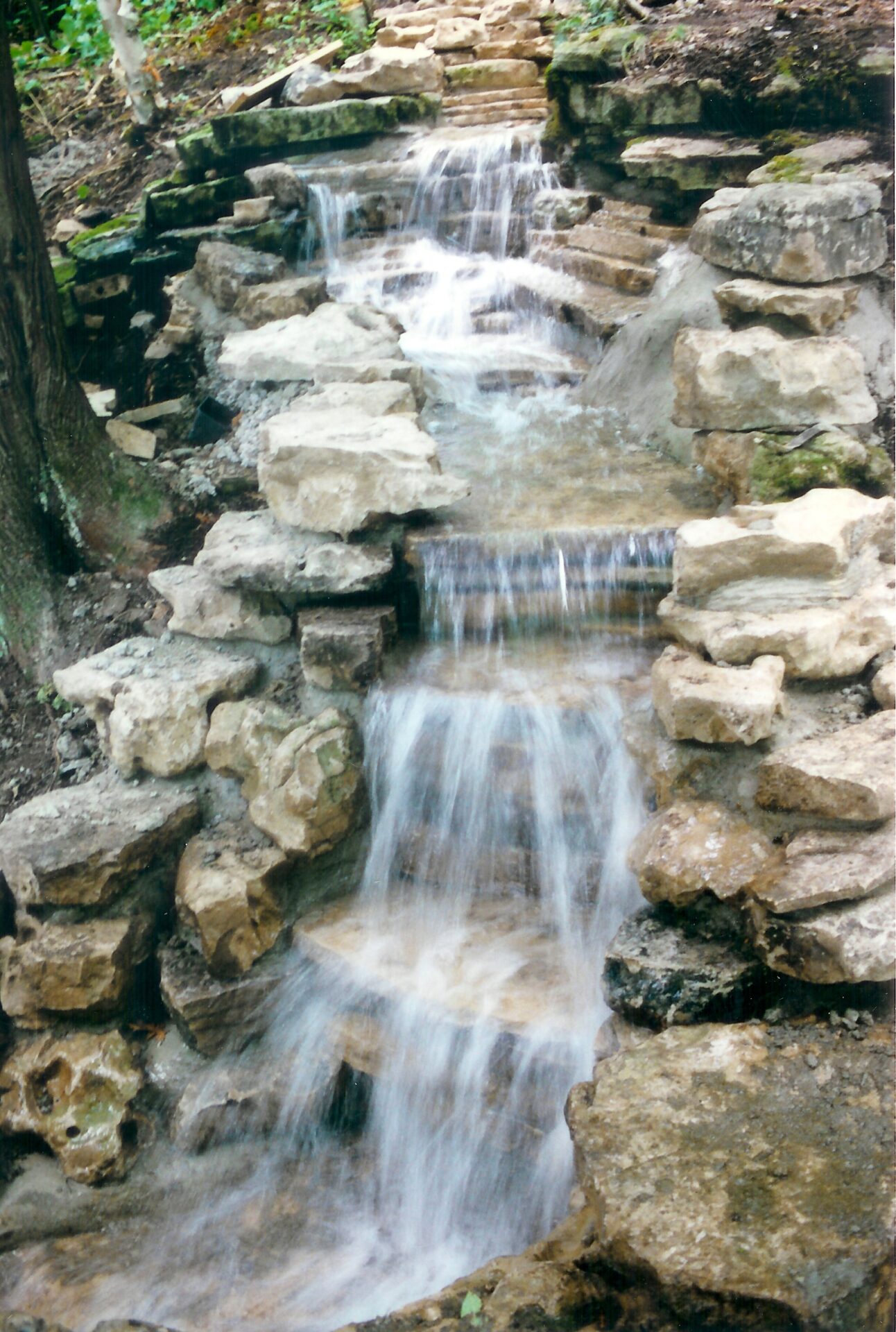 A small waterfall cascades over layered rocks in a forest setting, surrounded by greenery, creating a peaceful, natural landscape scene.