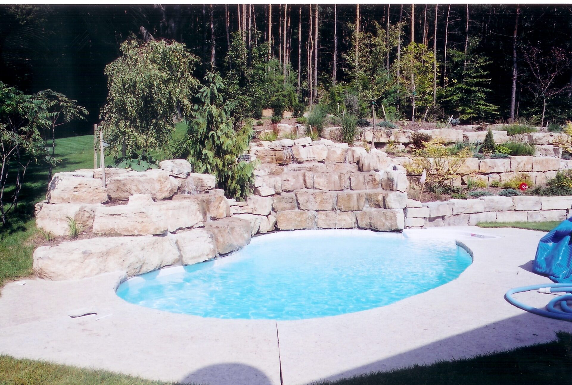 A small, kidney-shaped swimming pool with clear water is surrounded by stone walls and greenery, set against a backdrop of tall trees.