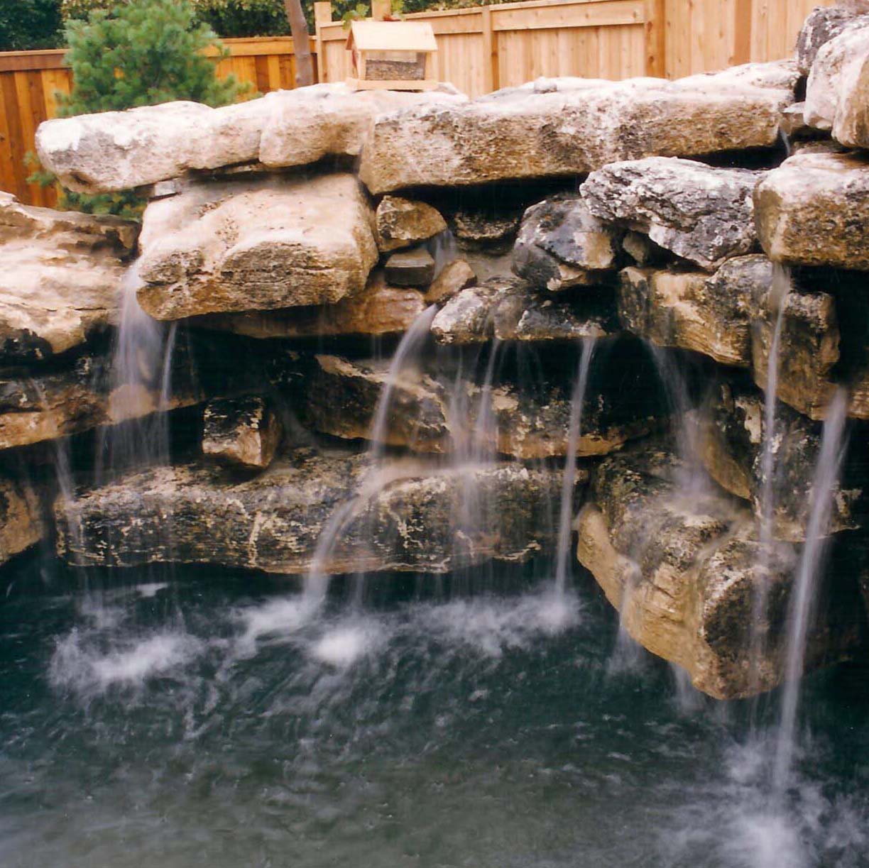 Natural stone waterfall feature with cascading streams into a small pond, set against a wooden fence and surrounded by greenery.