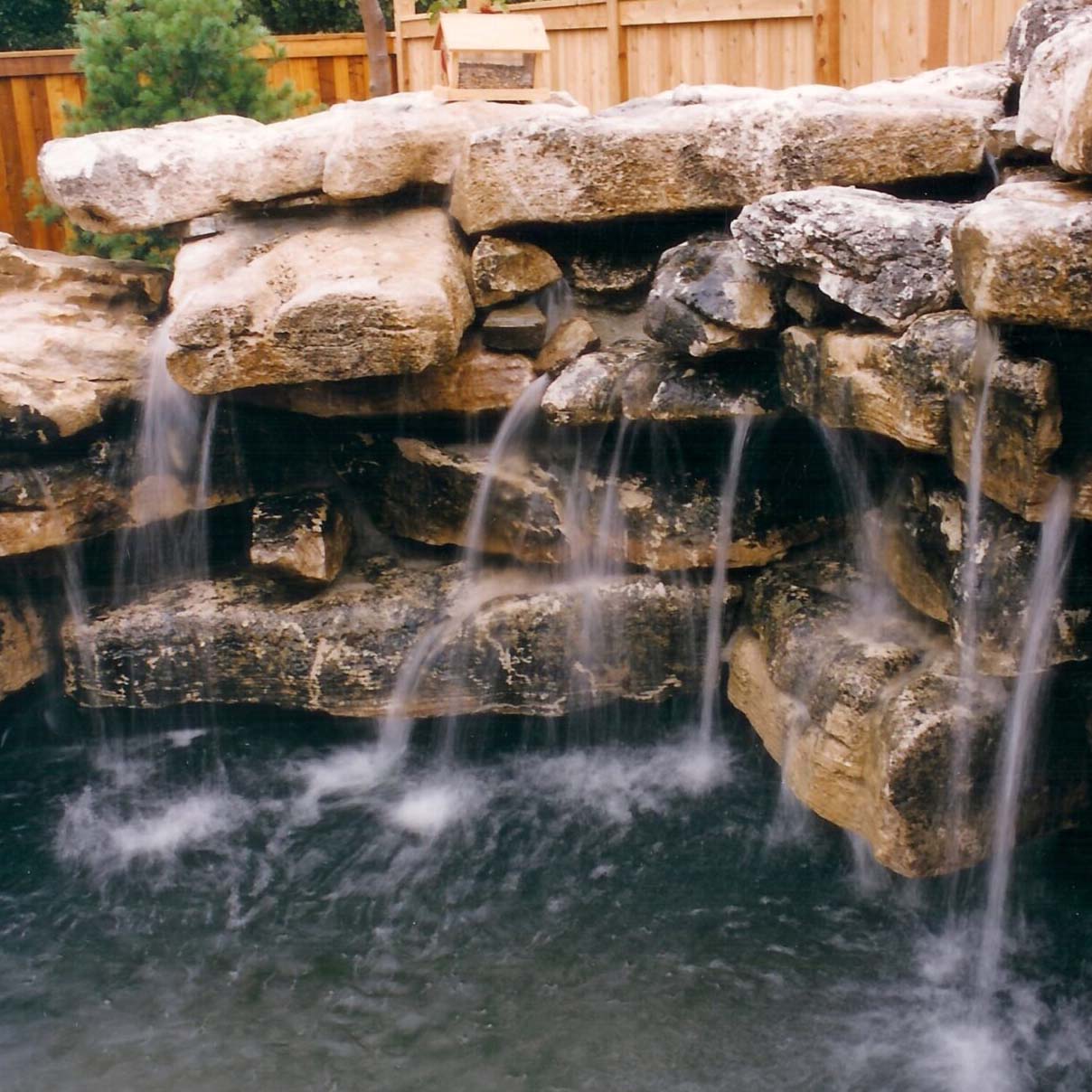 A small waterfall flows over a rocky ledge into a pond, enclosed by a wooden fence and surrounded by greenery.