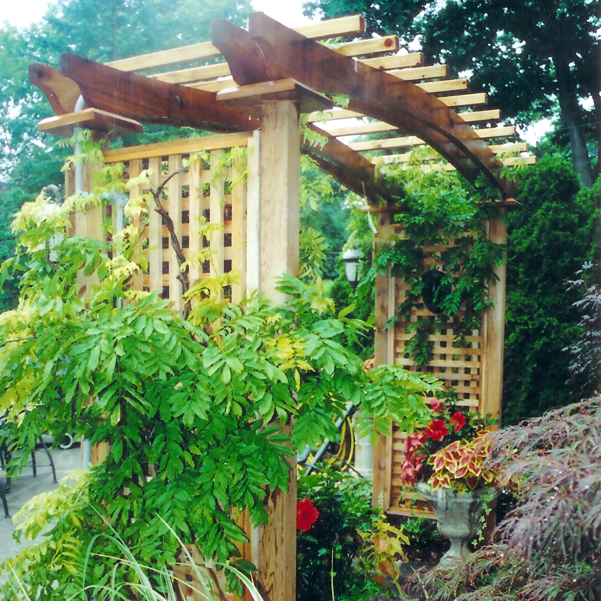 A wooden garden pergola covered in lush green vines and colorful flowers. Nearby is a lantern and patio seating area surrounded by foliage.