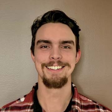 A person with a goatee and plaid shirt smiles against a plain background. The image focuses on the face, without additional context or landmarks.