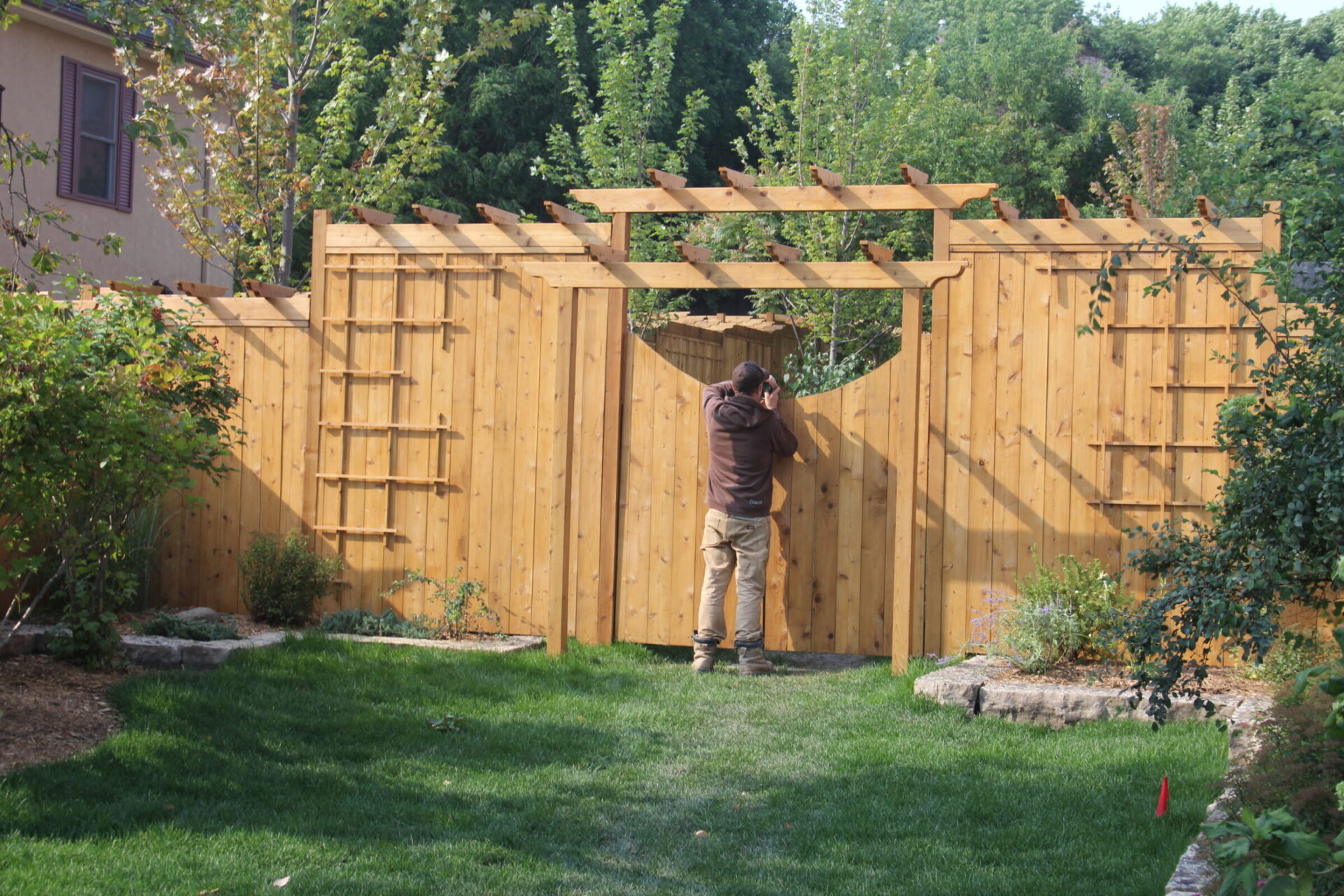 A person is inspecting a wooden fence in a garden. The fence features a gate with a circular decorative opening.