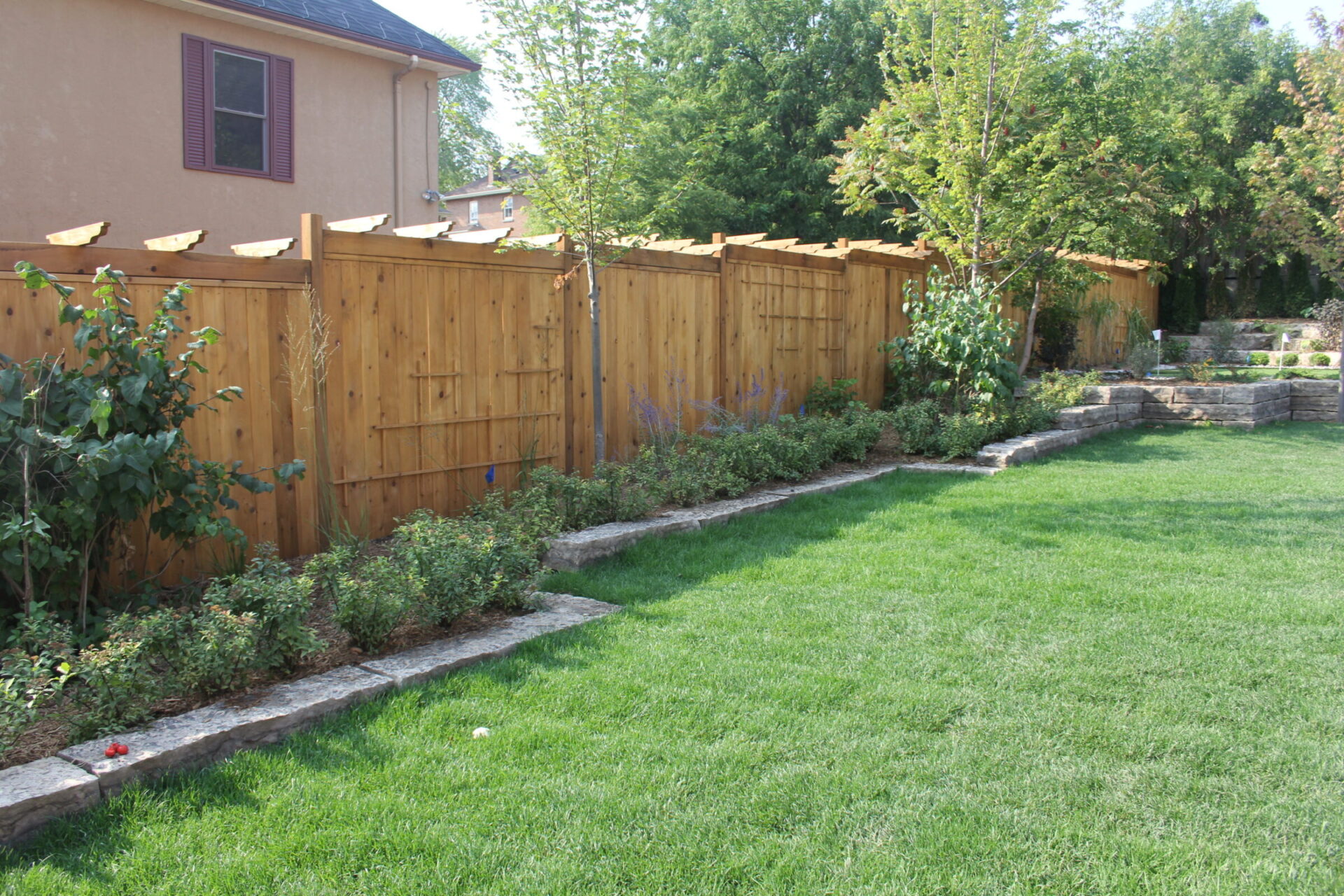 A backyard with green grass, a wooden fence, stone-bordered flowerbeds, and trees creating a relaxing, landscaped environment. No landmarks present.