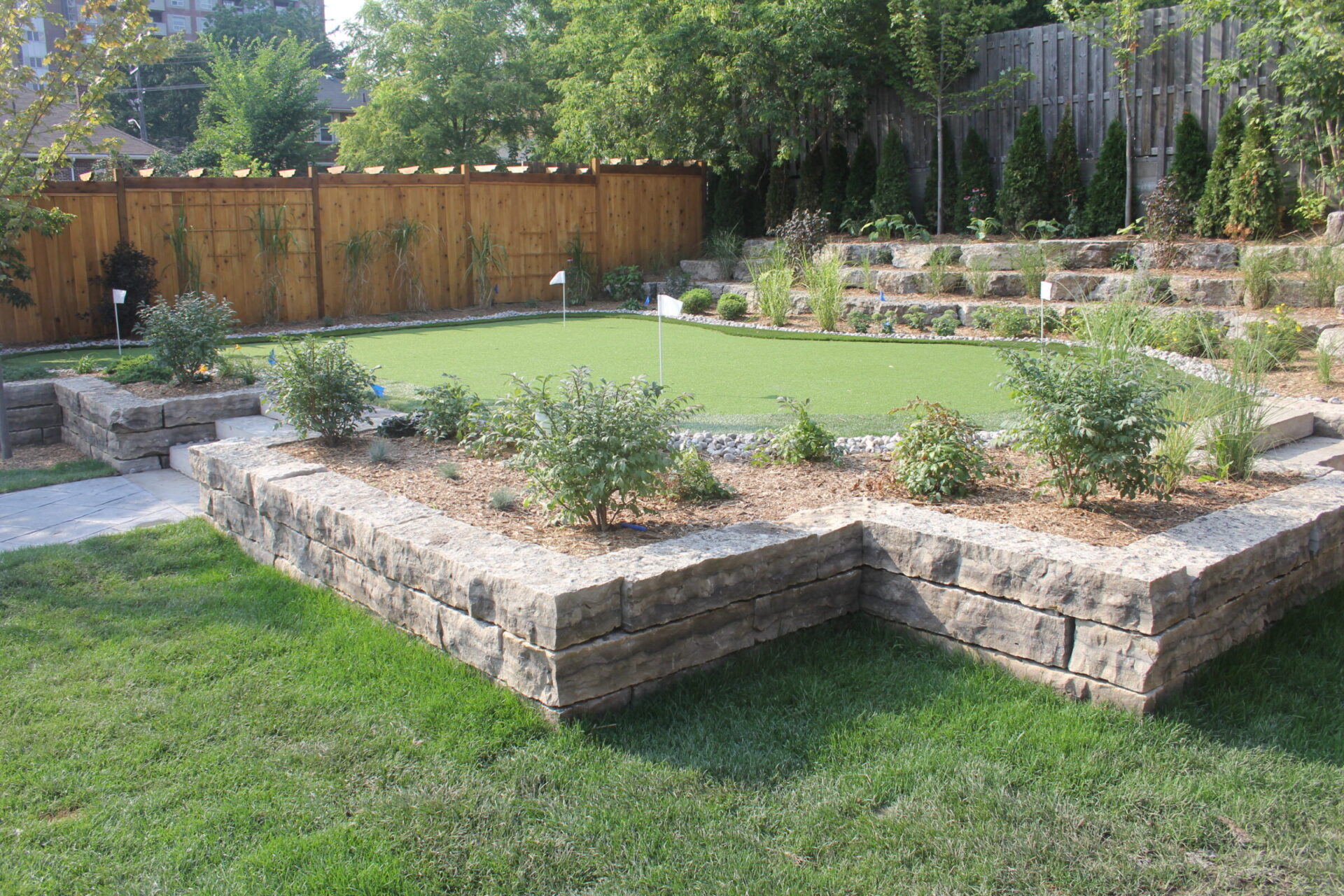 A neatly landscaped backyard features a stone-bordered putting green, surrounded by trees and shrubs, with a wooden fence in the background.