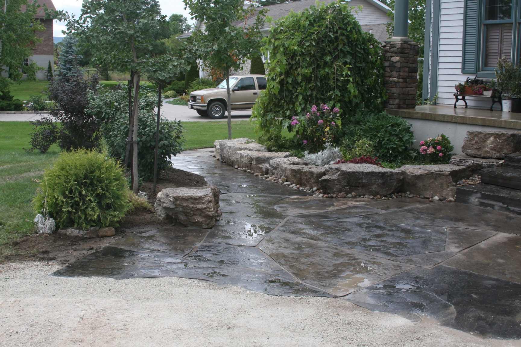 A garden pathway with stone steps, lush greenery, and flowering plants leads to the entrance of a house in a suburban neighborhood.