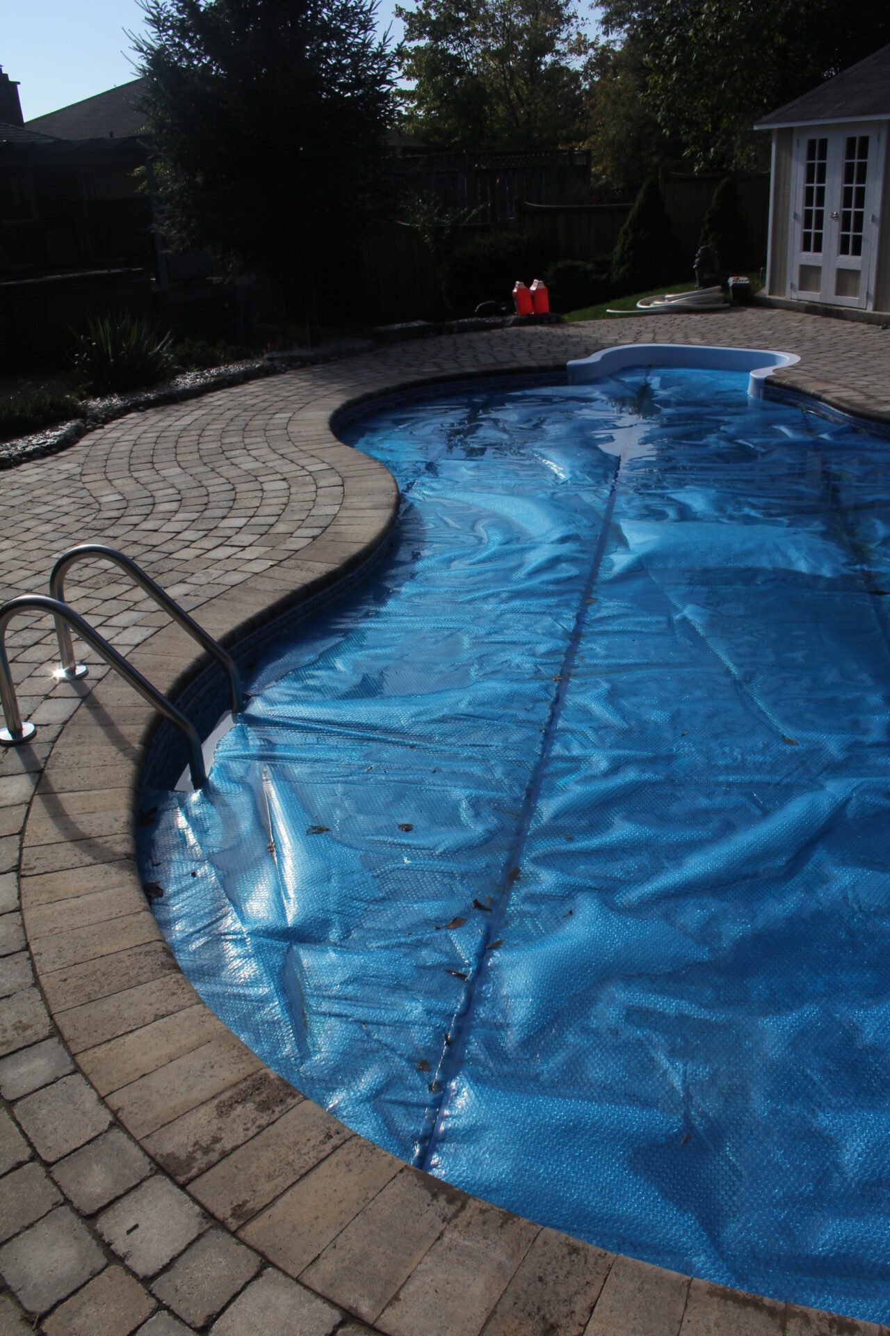 A backyard swimming pool covered with a blue tarp, surrounded by brick paving, trees, and a small white shed in the background.