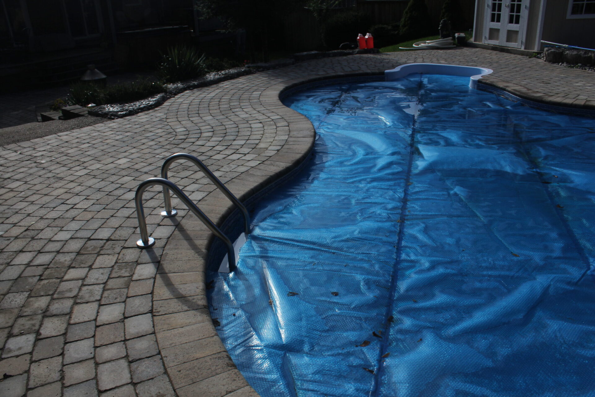 A backyard pool with a blue cover, surrounded by a stone patio. Ladders lead into the pool; no people are present.