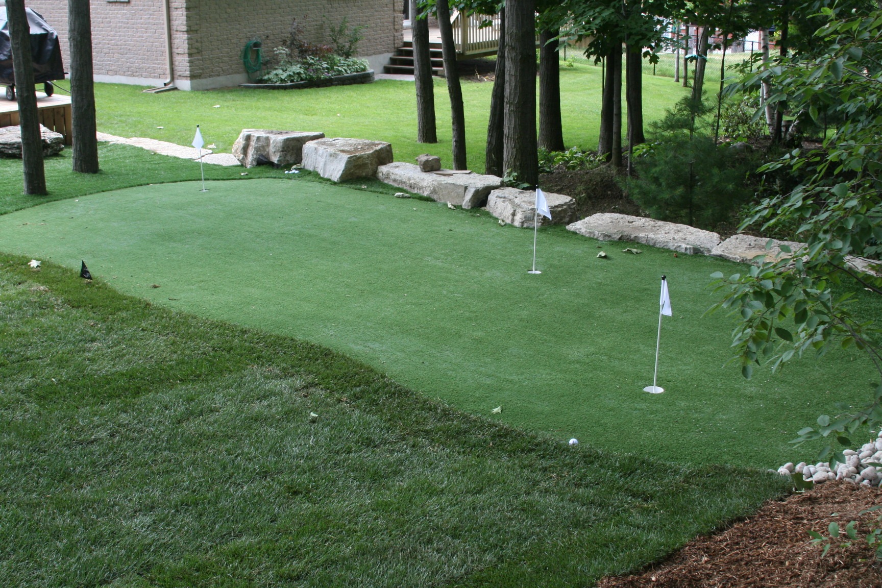 A small putting green with flags, surrounded by trees and rocks, is situated in a backyard with a nearby house.