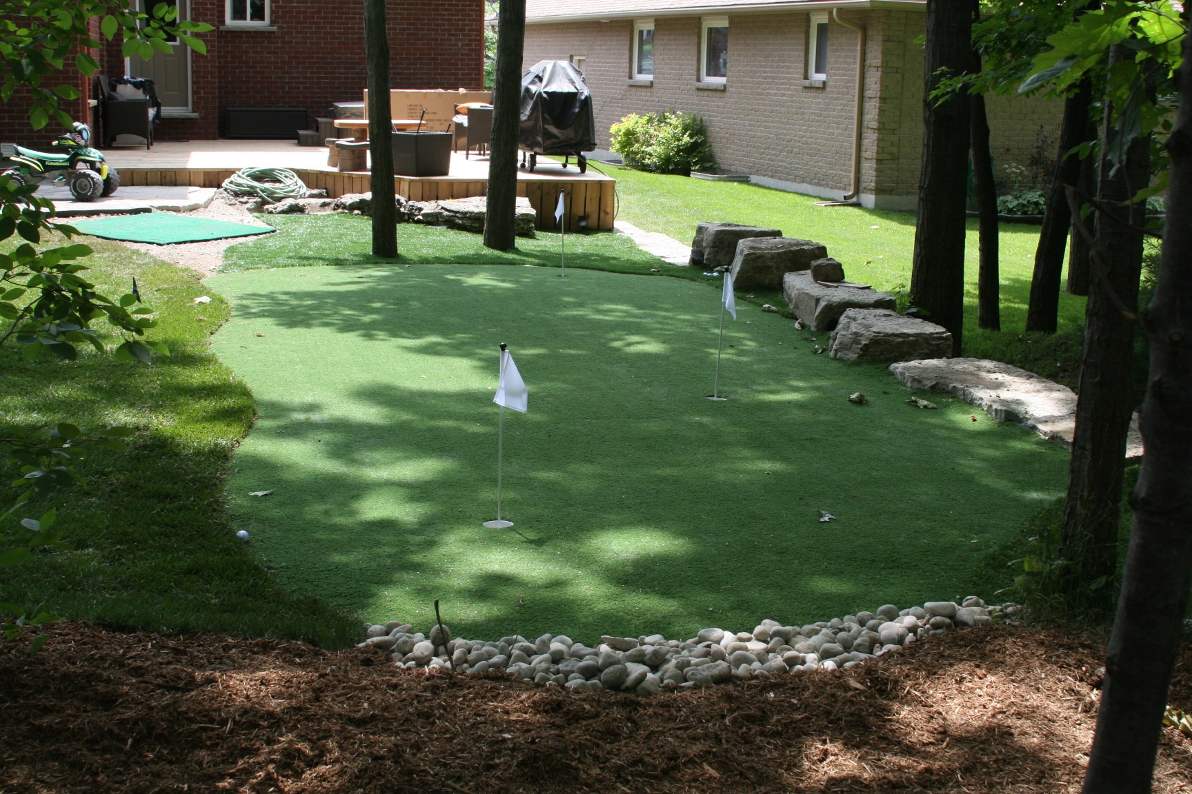 A backyard featuring a small putting green with flags, surrounded by stones and foliage, adjacent to a patio with chairs and a grill.