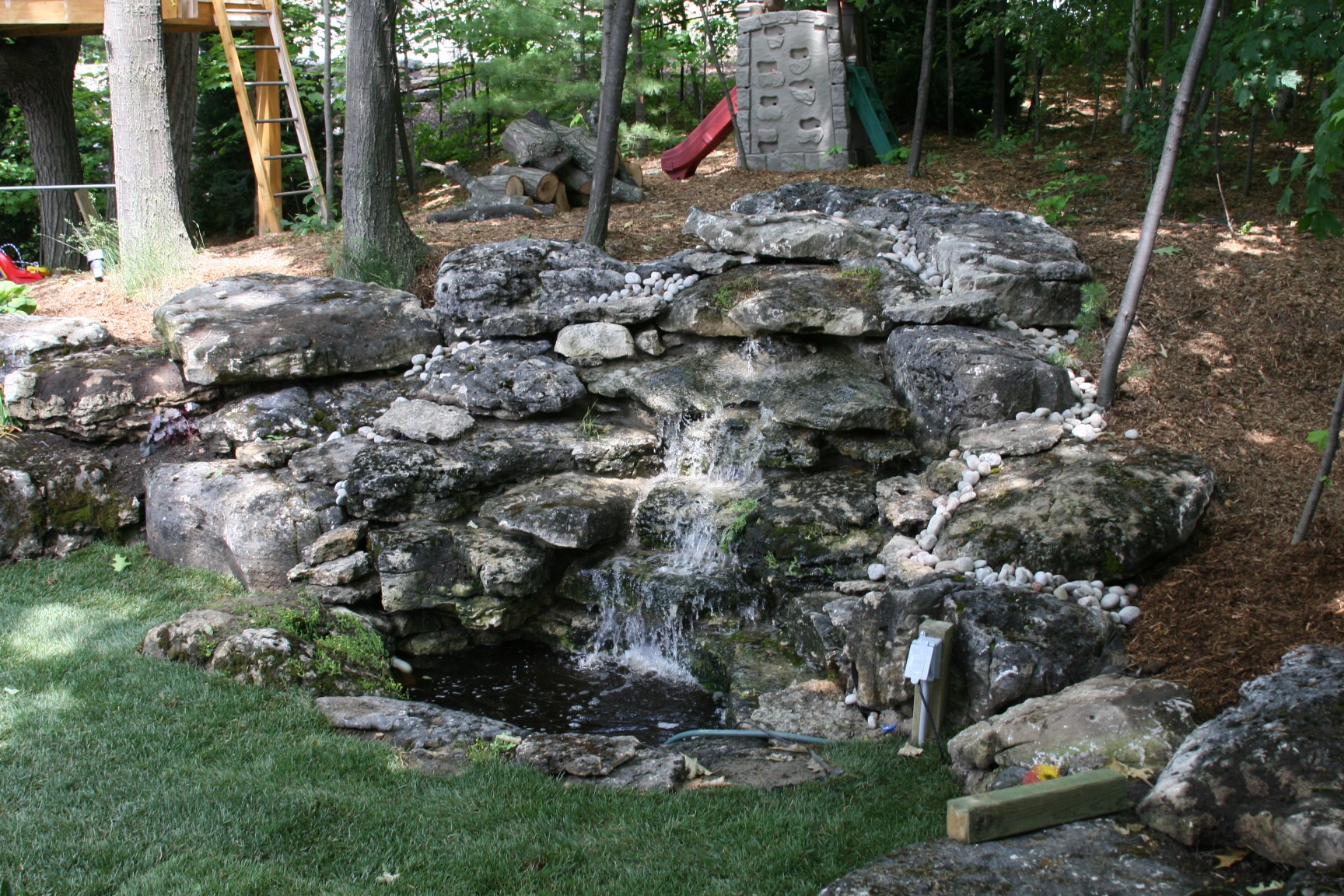 A small artificial waterfall flows over rocks in a backyard. Trees and a playground set are visible in the background.