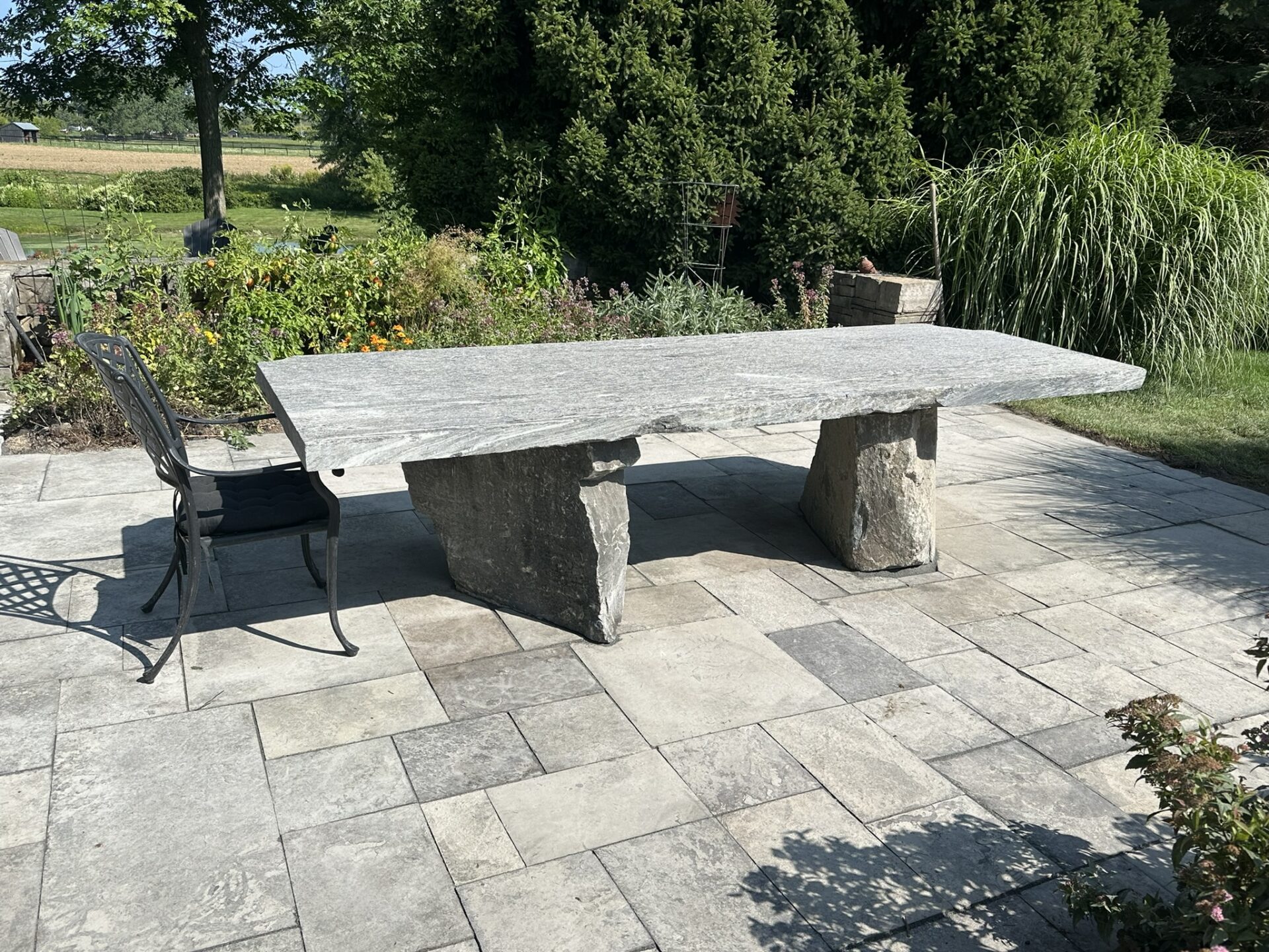 A stone table with a metal chair on a paved patio, surrounded by lush greenery and shrubs under a clear blue sky.