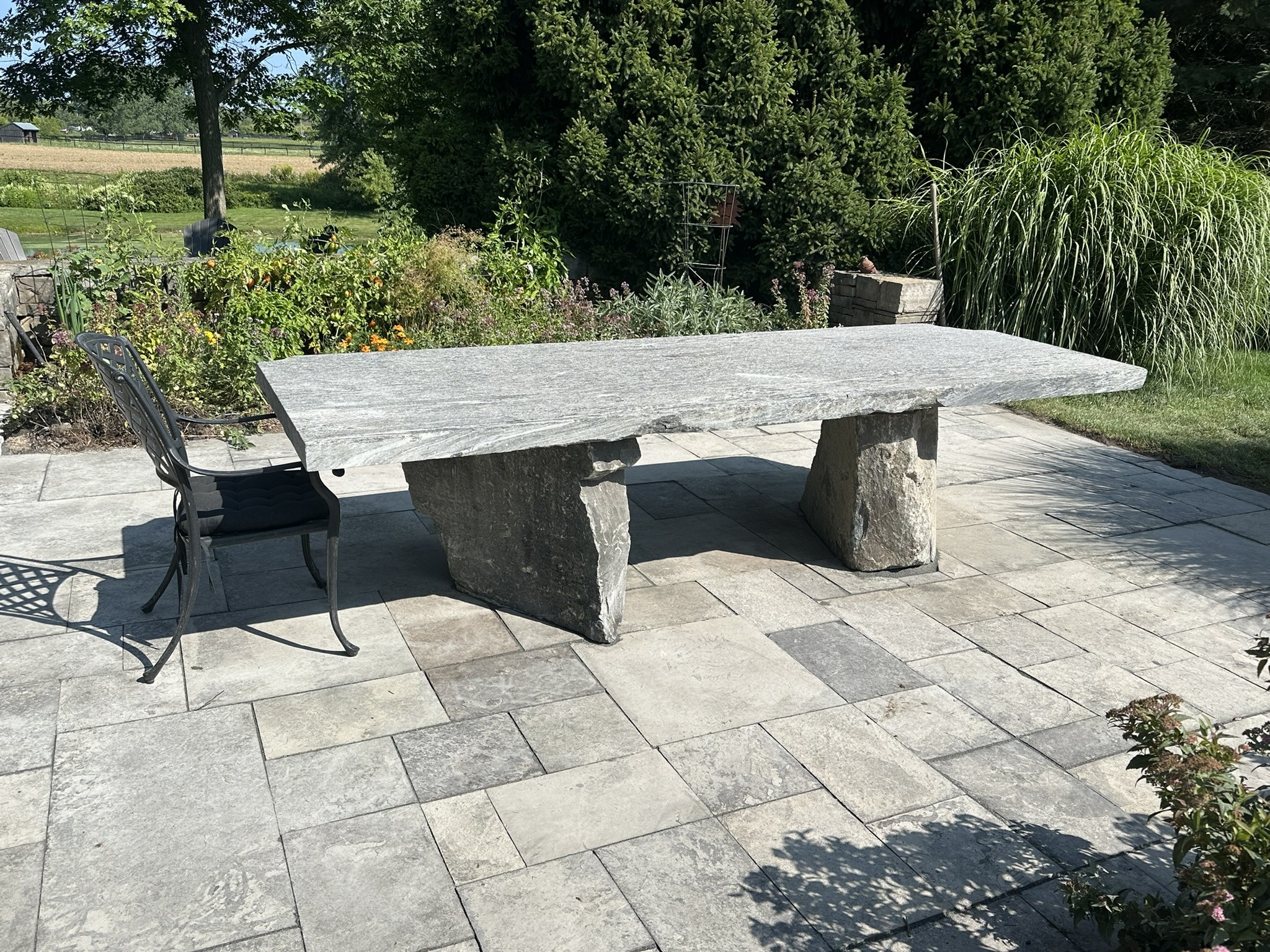 Stone table with metal chair on a paved patio, surrounded by lush greenery and garden plants under bright sunlight. No people or landmarks present.