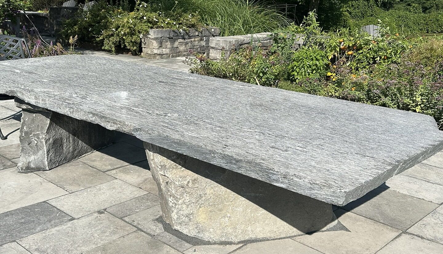 Stone table on a patio surrounded by greenery. Shadows cast on the ground. No recognizable landmarks or historical buildings visible.