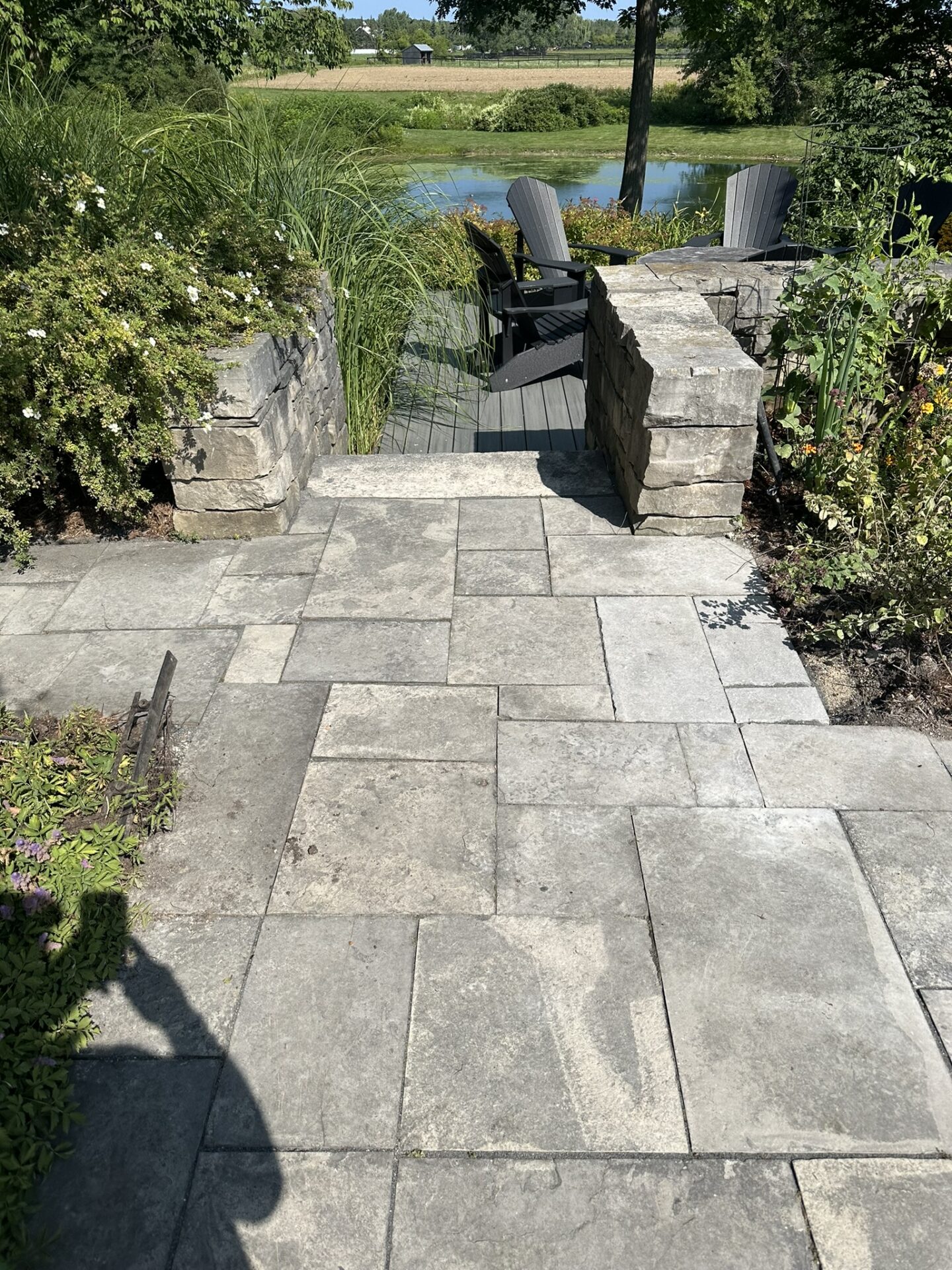 Stone pathway leads to a small patio with two chairs, overlooking a pond surrounded by lush greenery and trees under a clear sky.