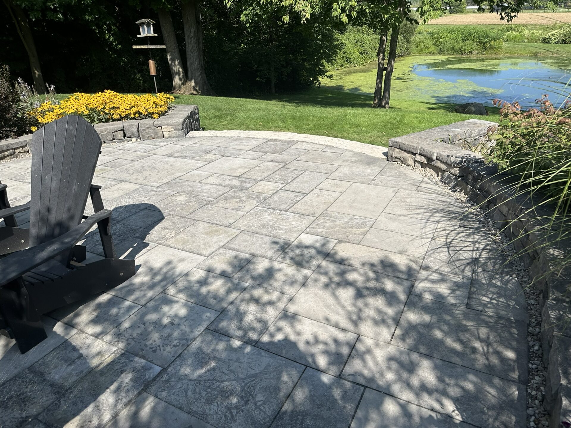 Stone patio with black Adirondack chair, overlooking a tranquil garden pond. Surrounded by lush greenery and bright yellow flowers under clear skies.